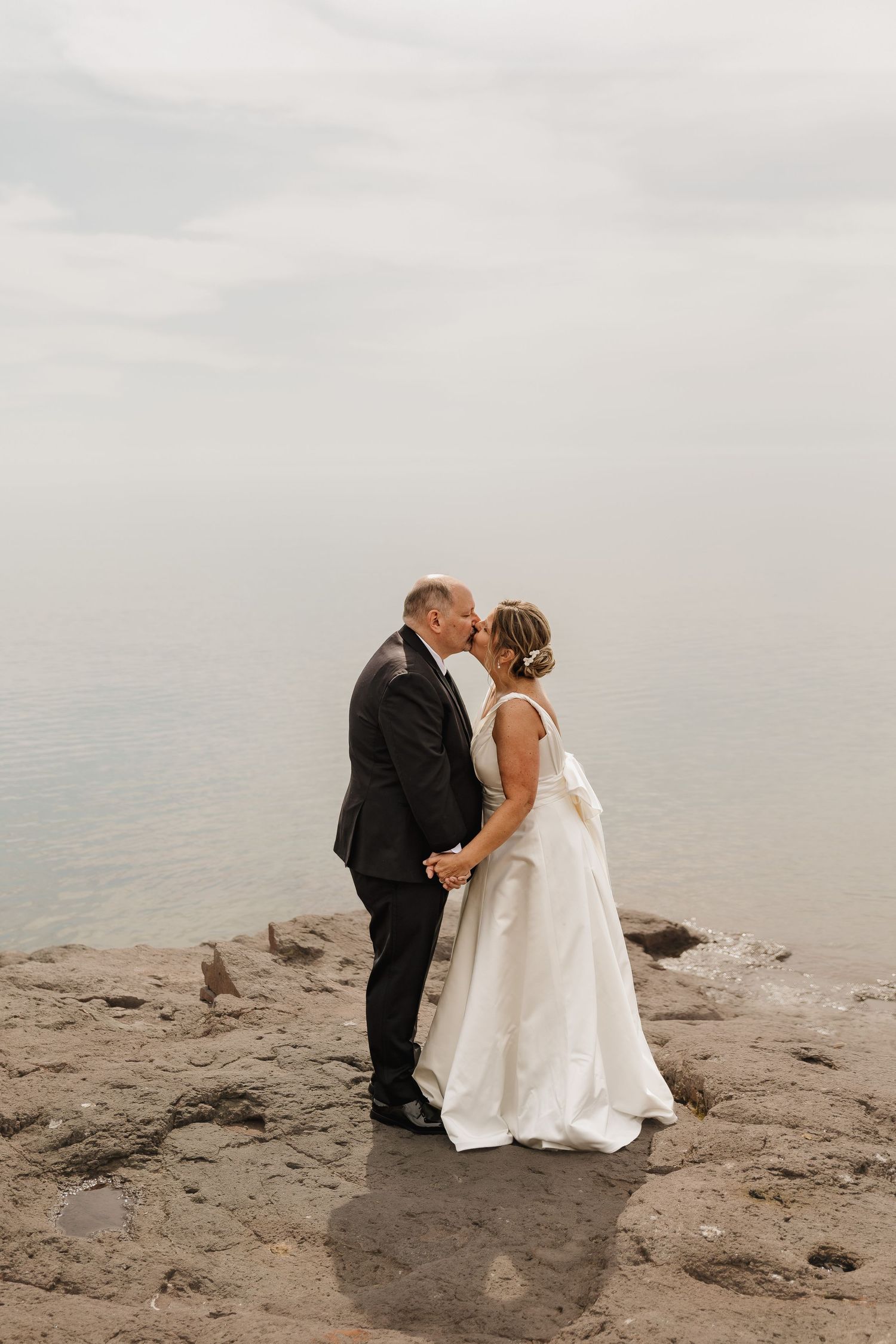 Bride and groom kissing on rocky shoreline in wedding attire with calm ocean and overcast sky backdrop.