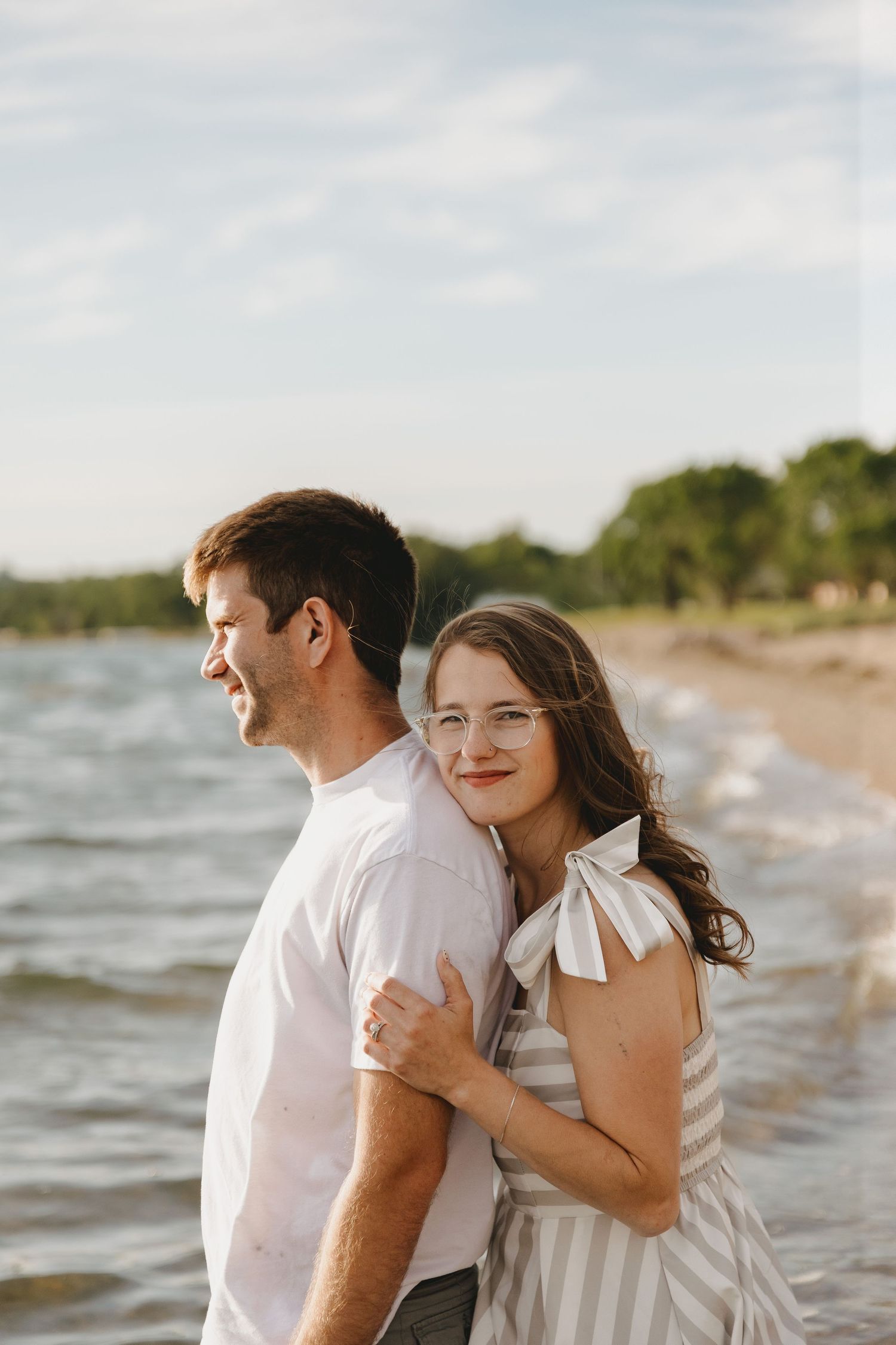 A couple embraces on a sandy beach at golden hour, woman in striped dress smiling at camera, man in white shirt.