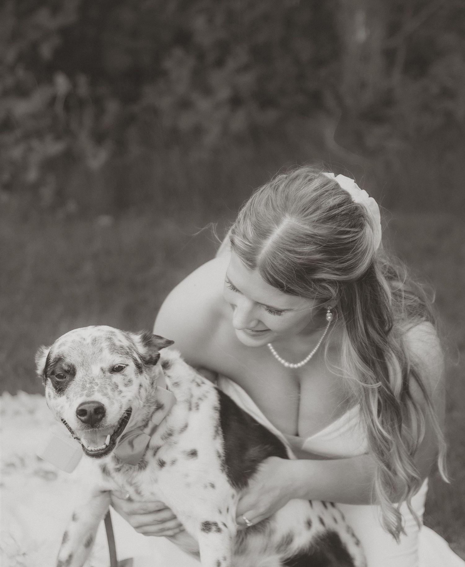 Black and white photo of a smiling bride in a strapless gown hugging her spotted dog outdoors.