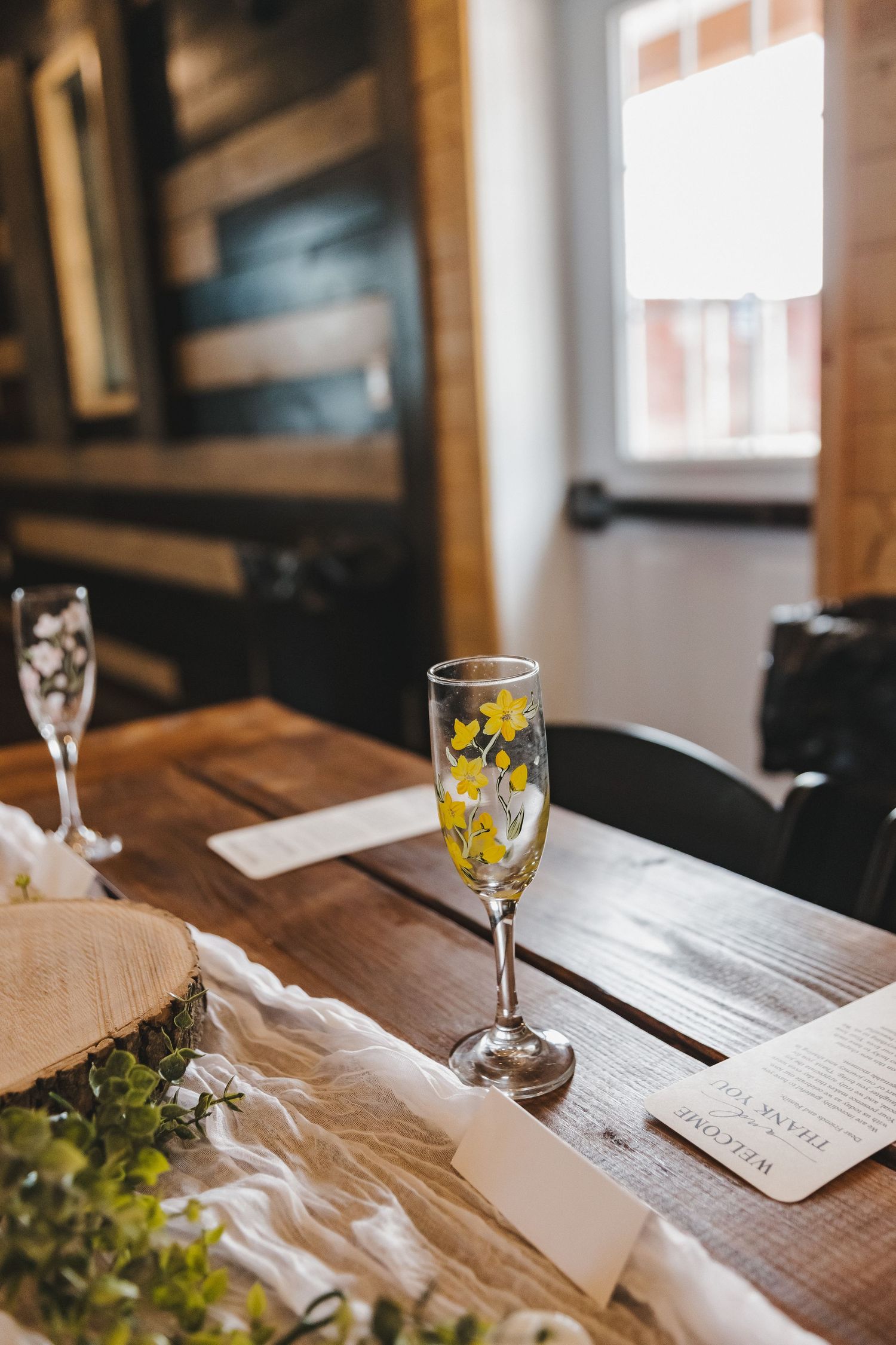 Elegant champagne flutes with yellow floral details on a rustic wooden wedding reception table with greenery.
