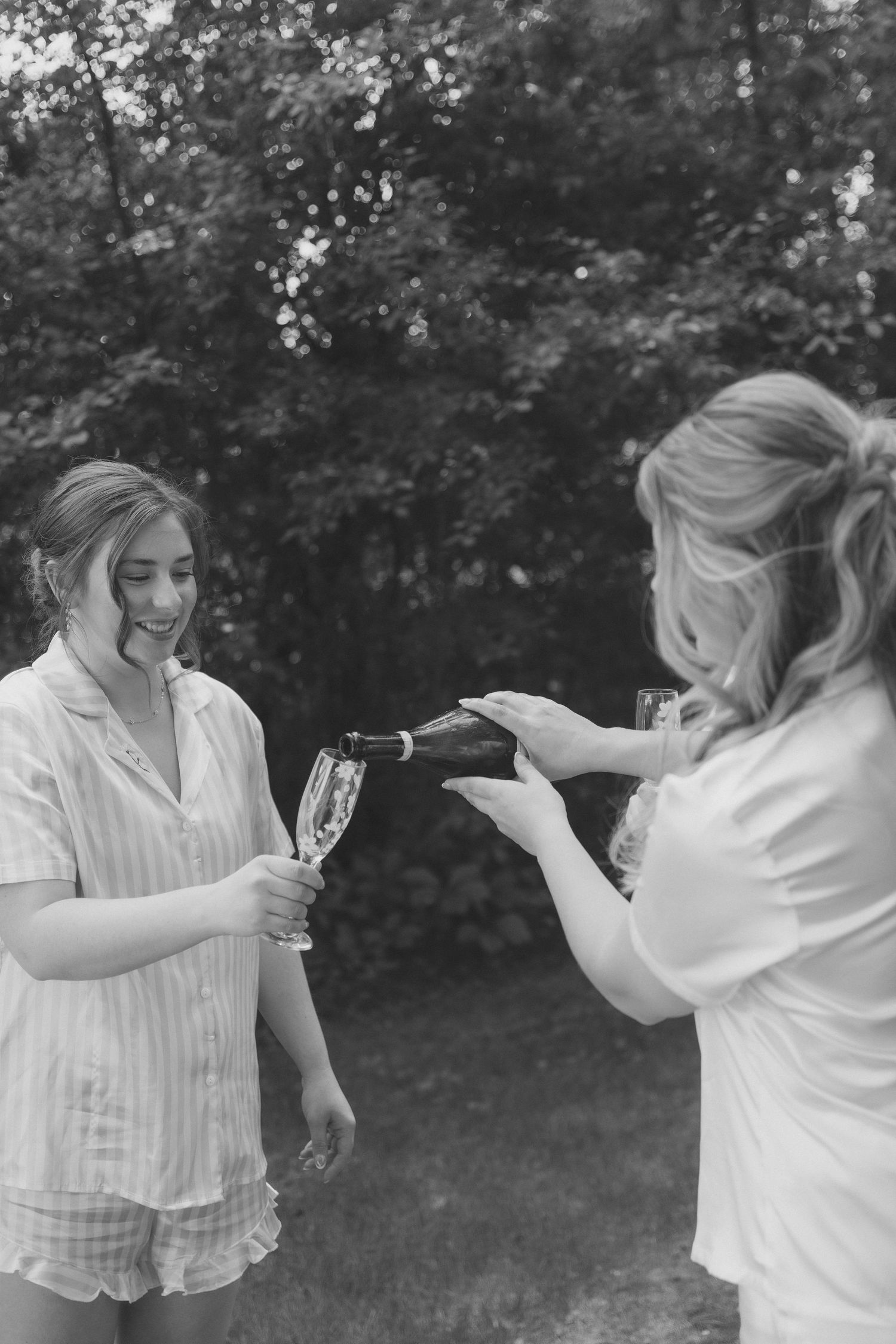 Two women in pajamas pouring champagne outdoors in a black and white photo.