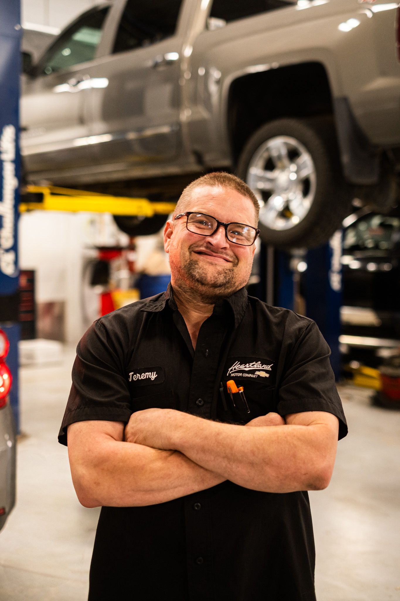 Service professional in black uniform smiling in auto repair shop with truck on lift behind.