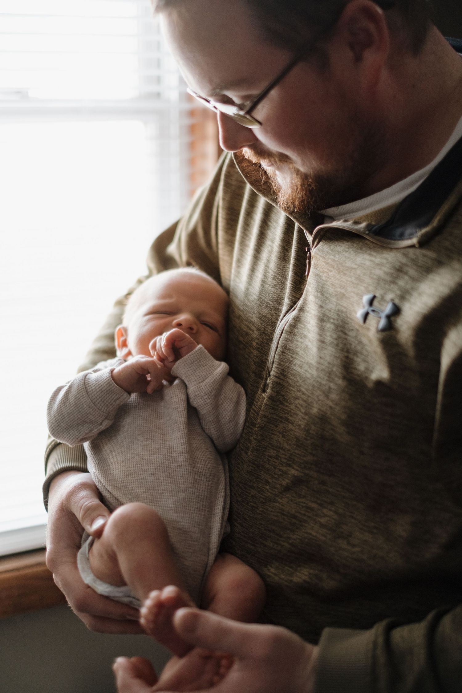 Parent in glasses holding peaceful sleeping infant in gray onesie by window.