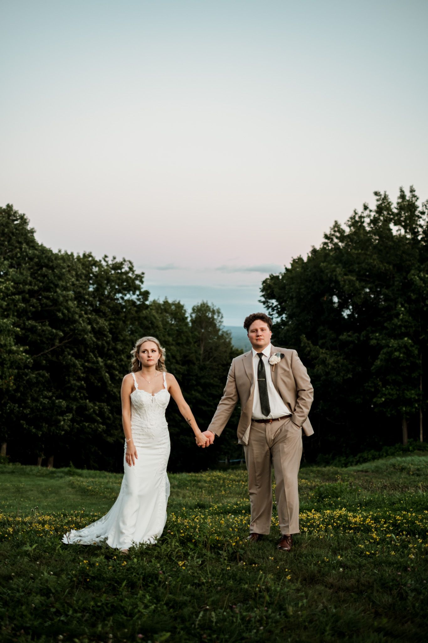 Newlyweds holding hands while walking through wildflower field at sunset.