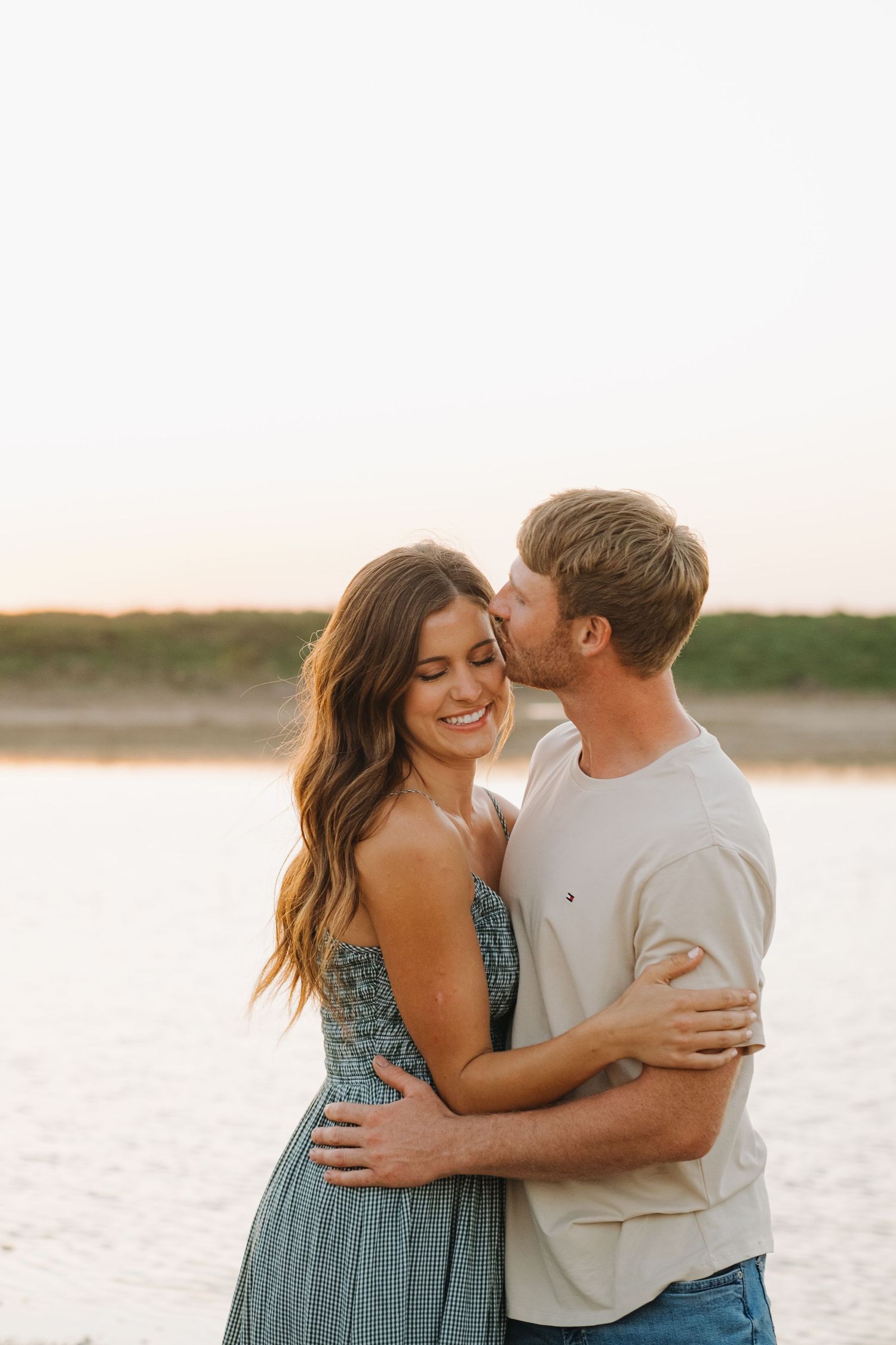 Romantic couple embraces on a beach at sunset wearing casual summer outfits.