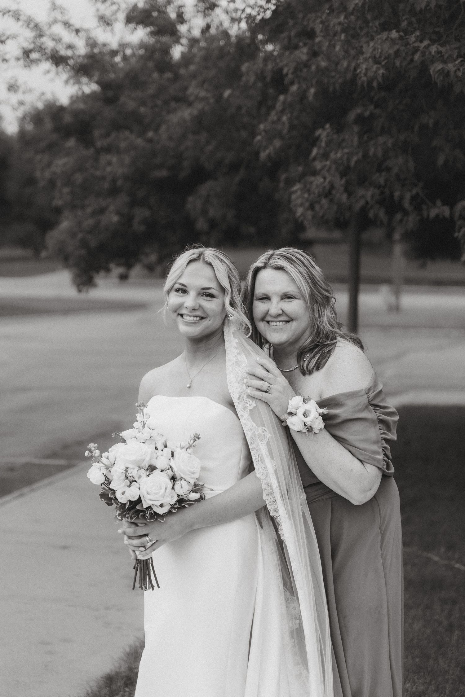 Bride holding white rose bouquet smiles with older woman embracing her in black and white wedding portrait.
