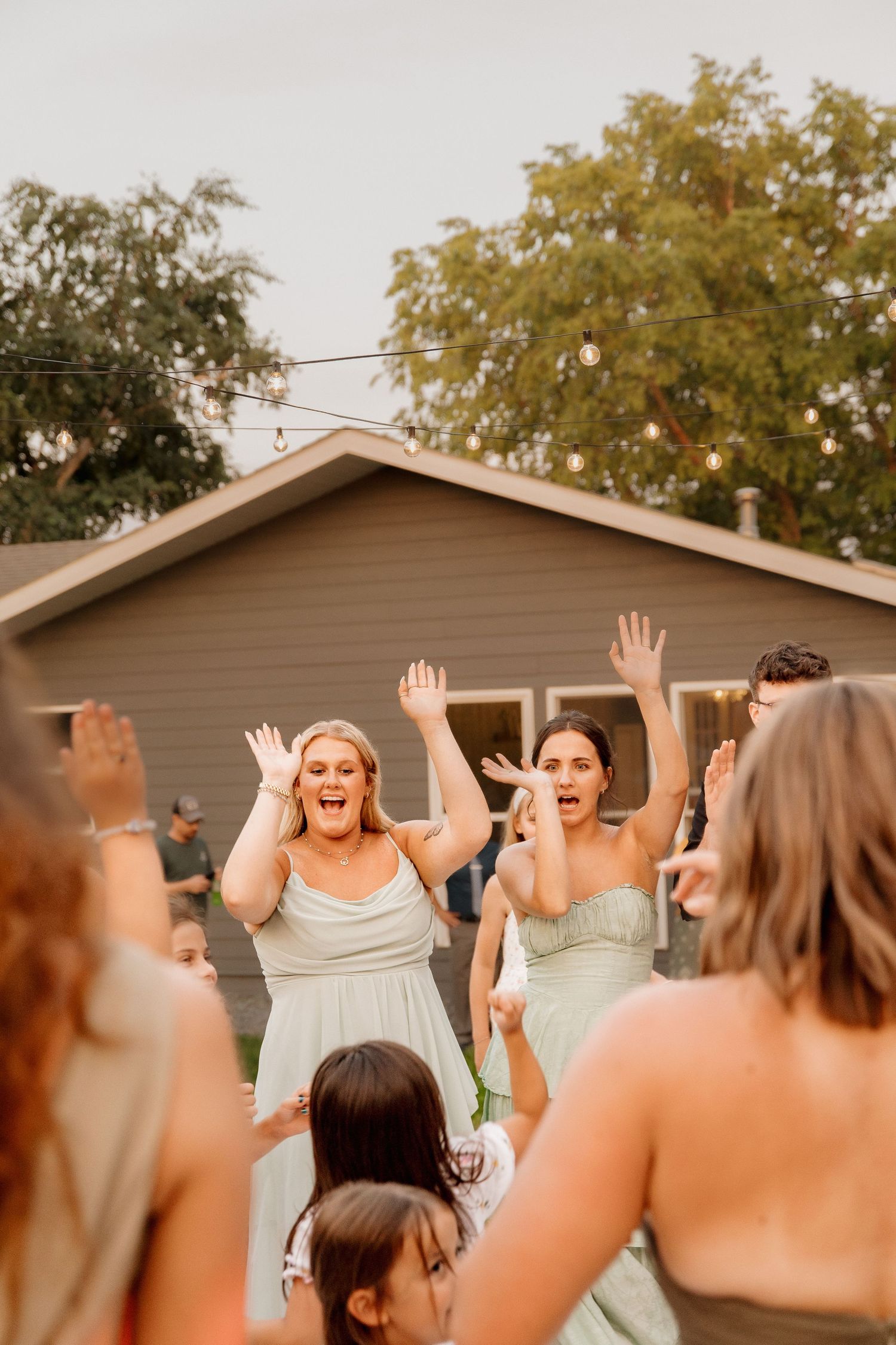 Excited bridesmaids in sage green dresses dancing with hands raised at outdoor wedding reception.