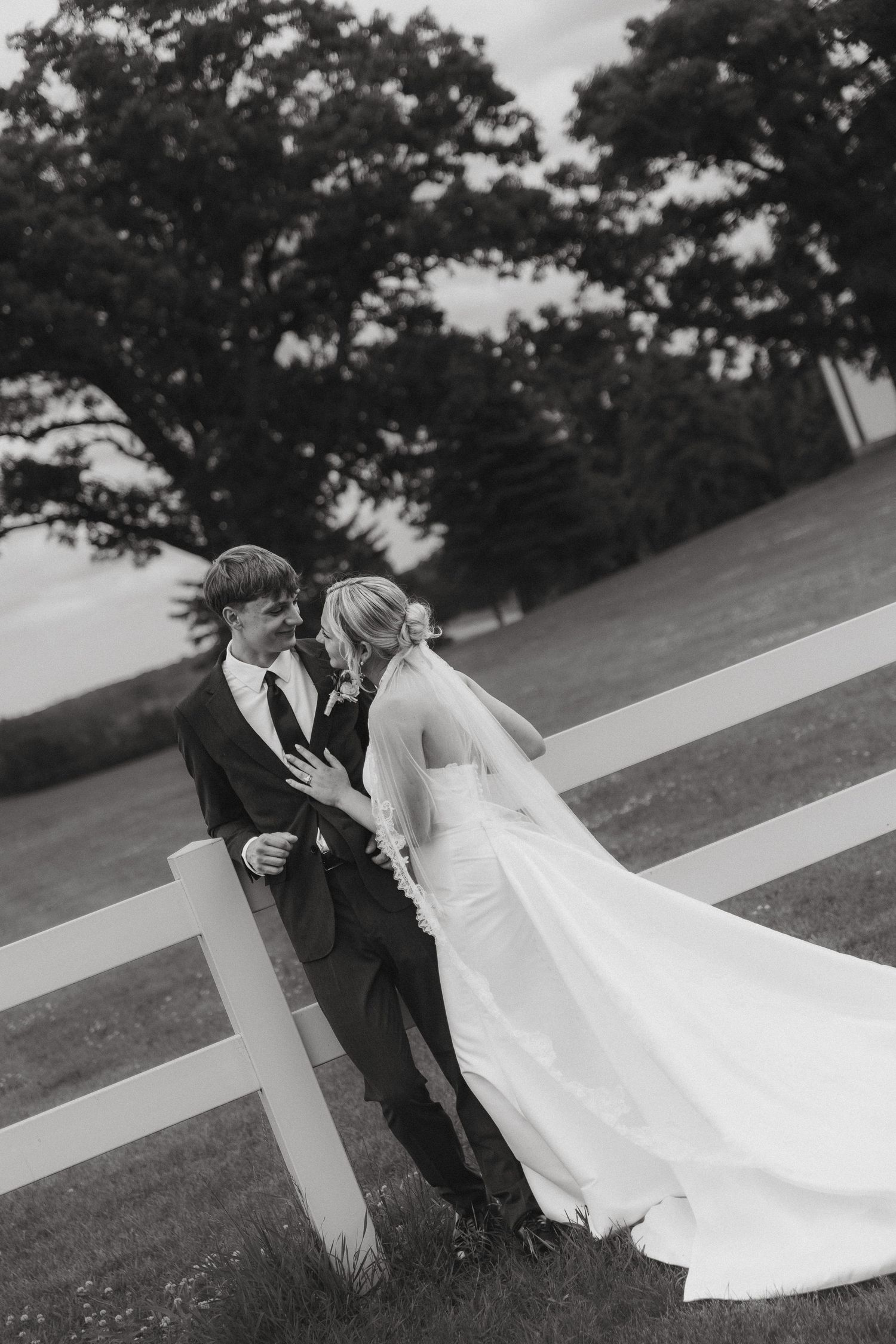 Bride and groom share an intimate moment leaning against a white fence in a black and white wedding photo.