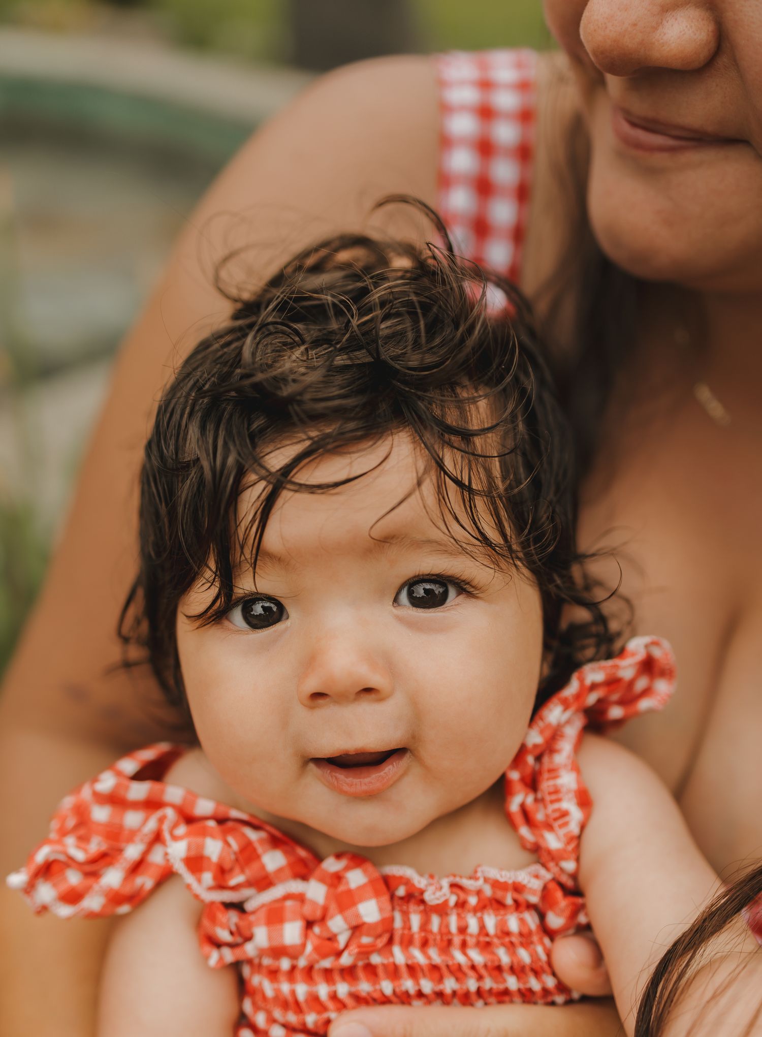 Adorable baby with dark curly hair in red gingham outfit held by mother outdoors.