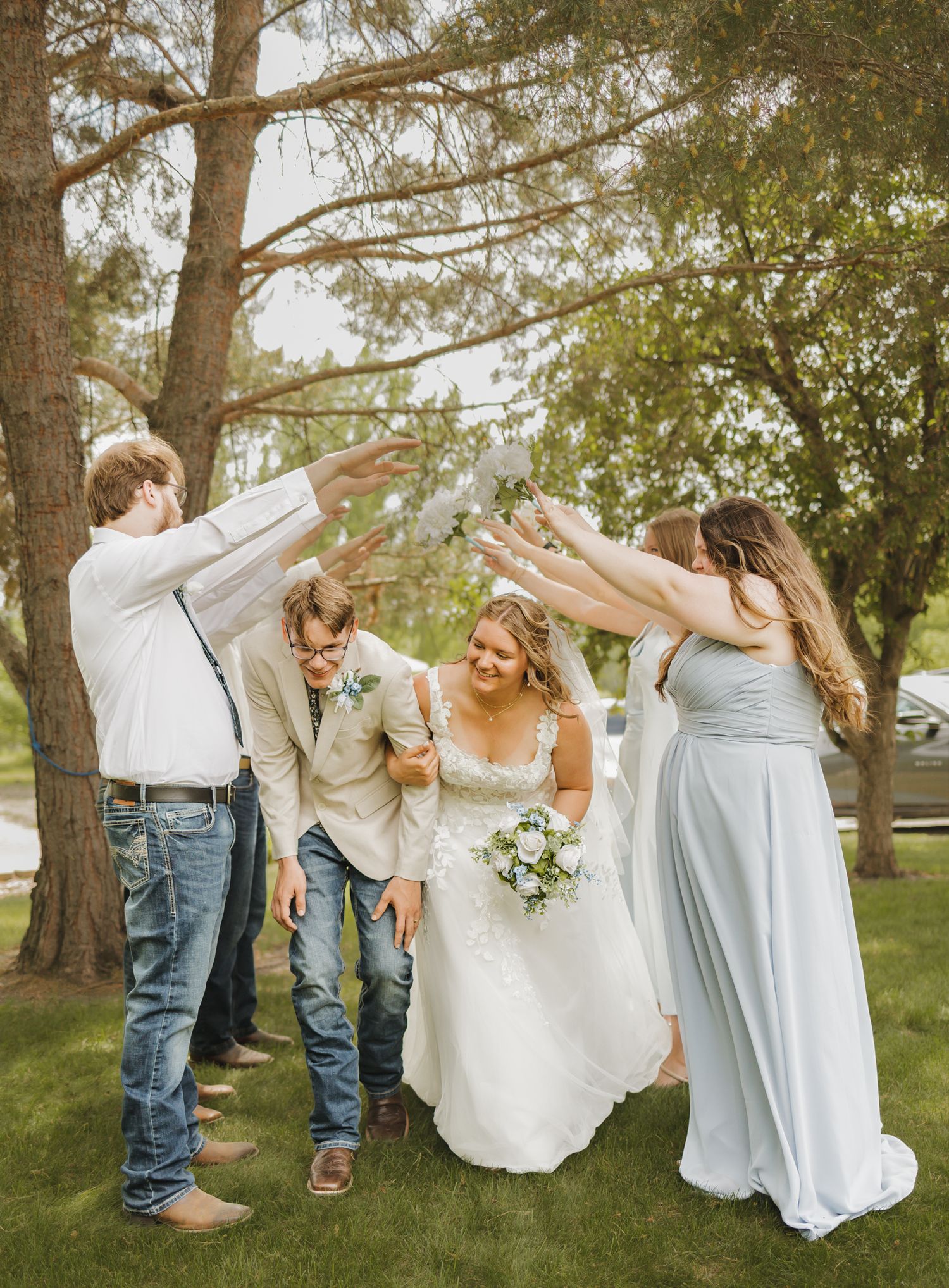 Wedding party celebrating outdoors, bride in white gown holding bouquet, bridesmaids in blue, groomsmen raising hands.