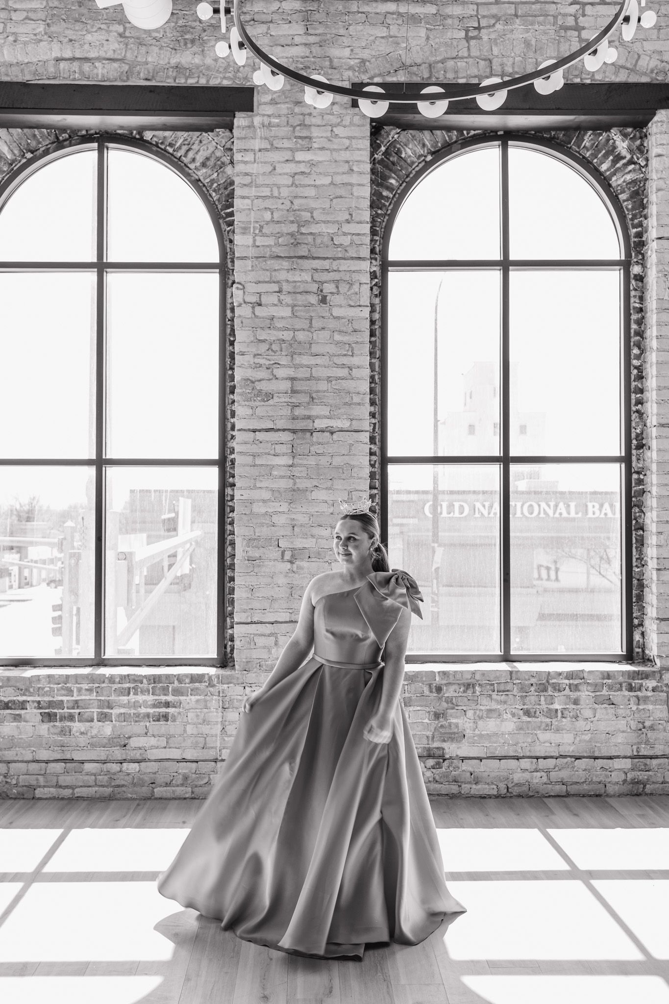 Woman in flowing ball gown poses gracefully in sunlit industrial loft with arched windows and brick walls.