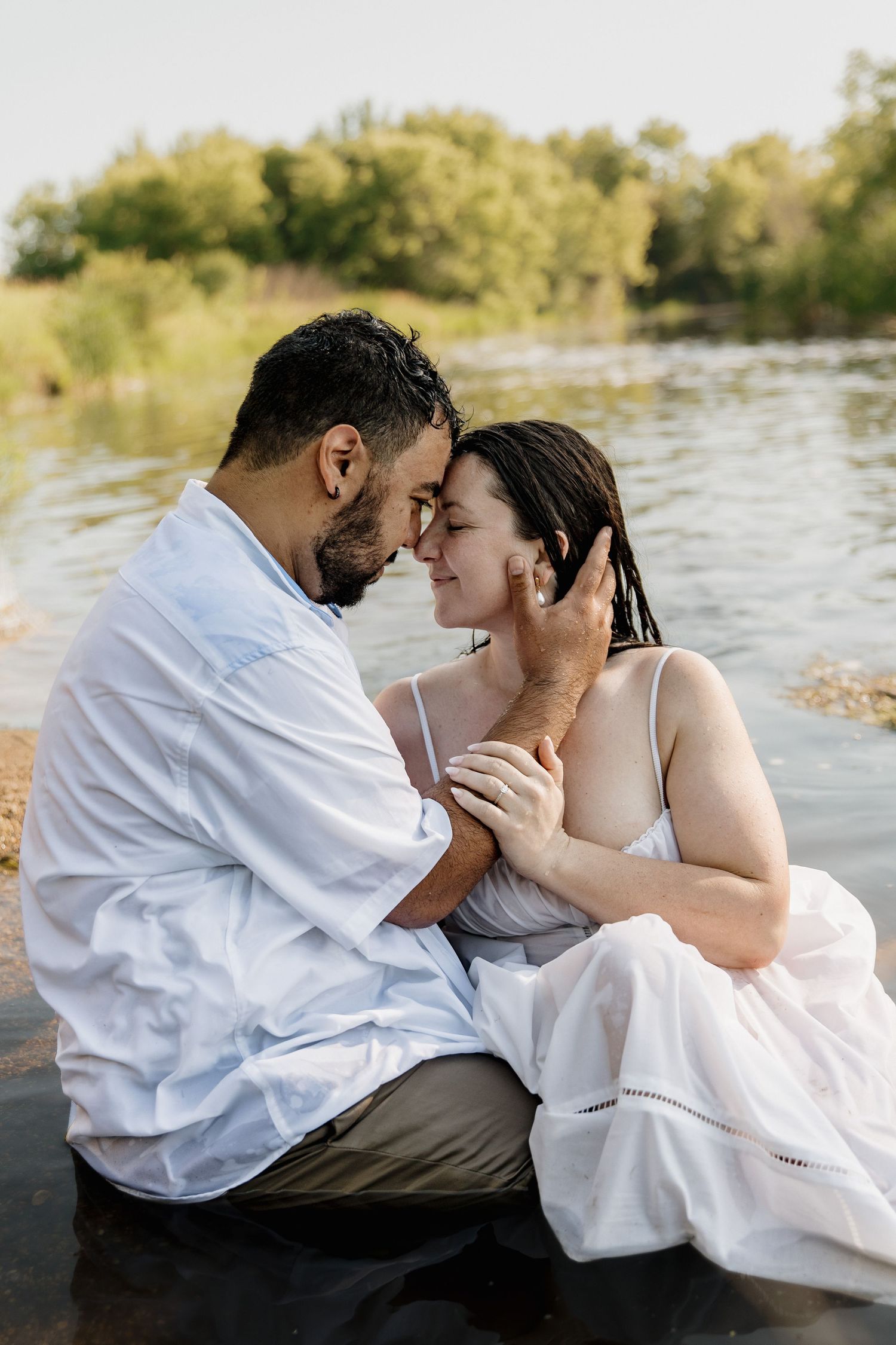 Romantic couple sitting by a serene river, sharing an intimate moment in white outfits surrounded by lush greenery.