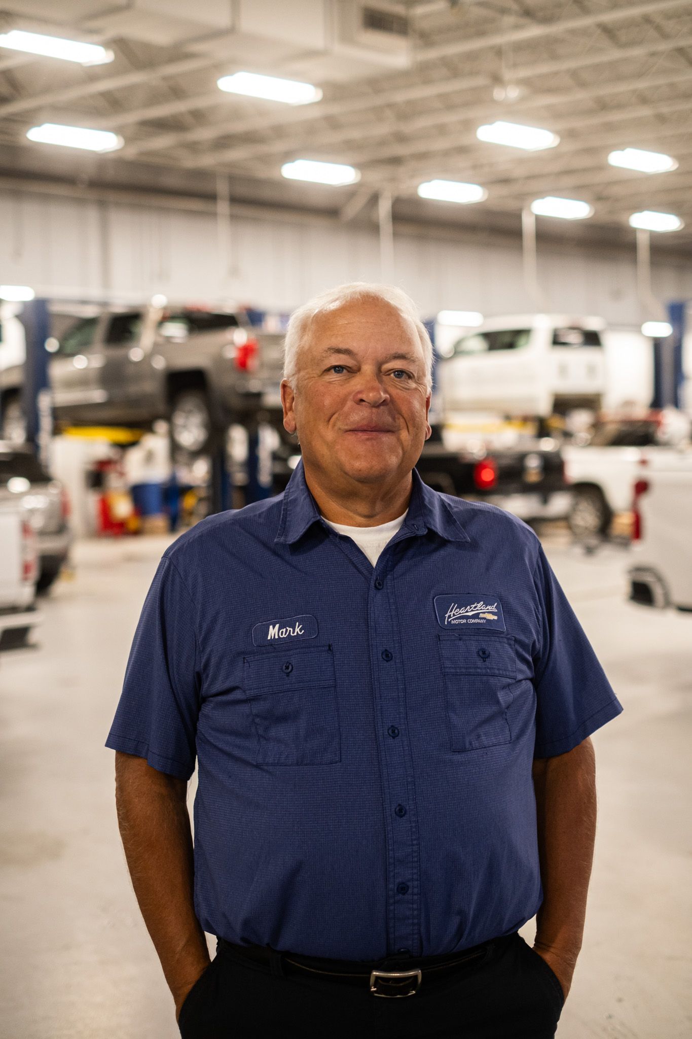 Auto service professional in navy uniform standing in large repair facility with vehicles on lifts.