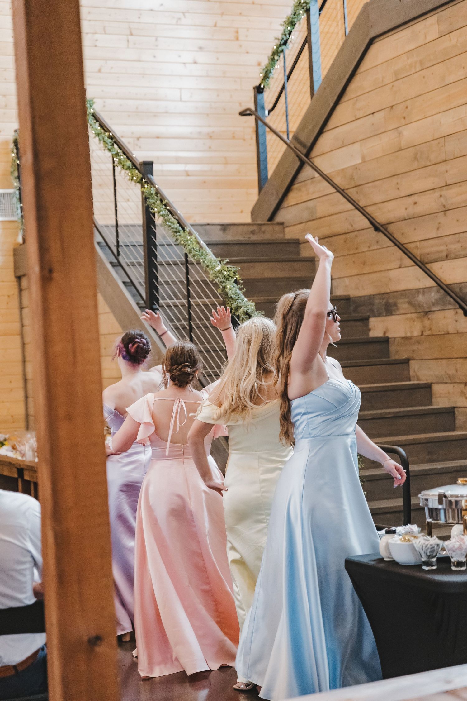 Bridesmaids in colorful gowns reaching up near staircase at wedding venue.