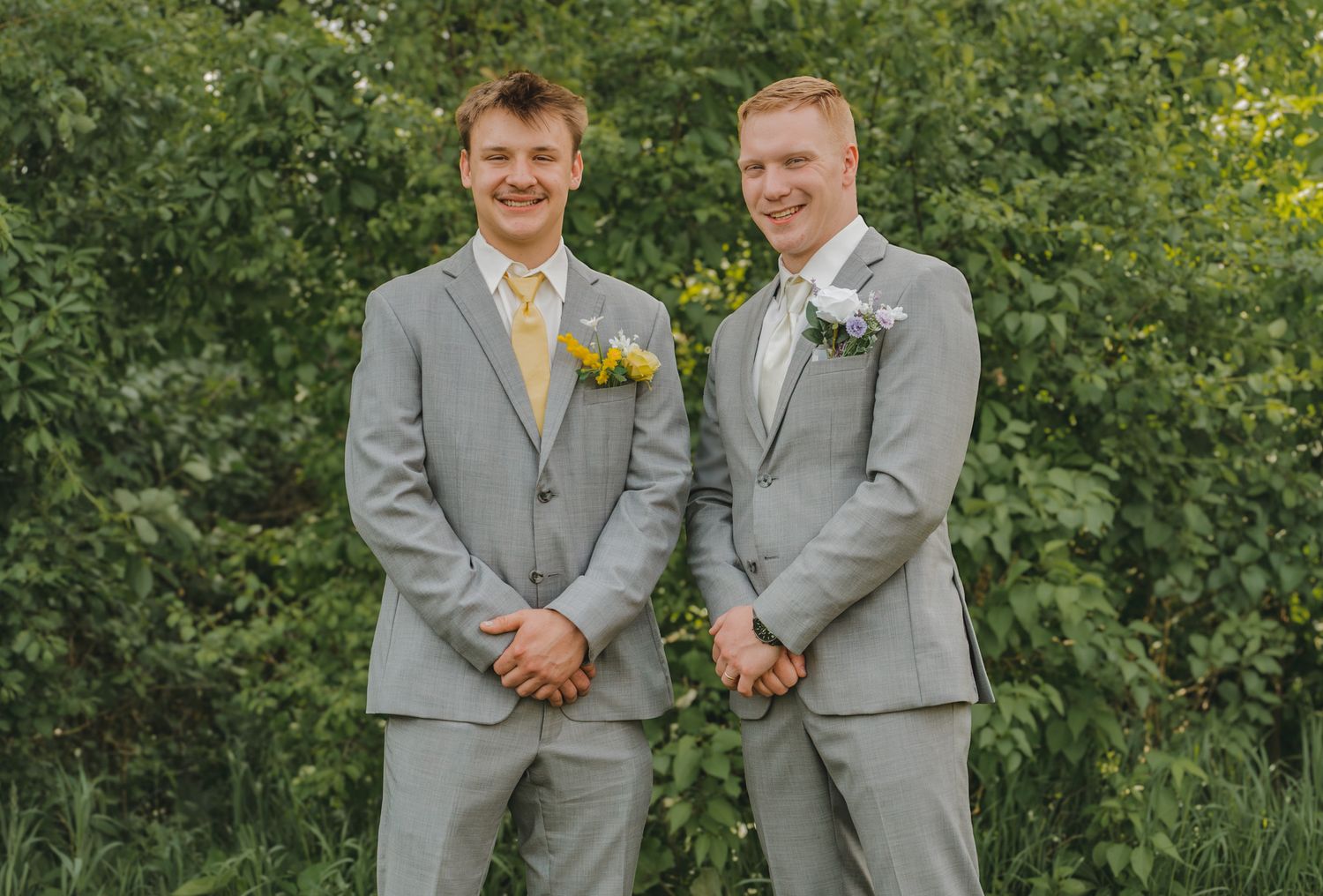 Two young men in matching gray suits with boutonnieres smiling outdoors at a wedding ceremony.