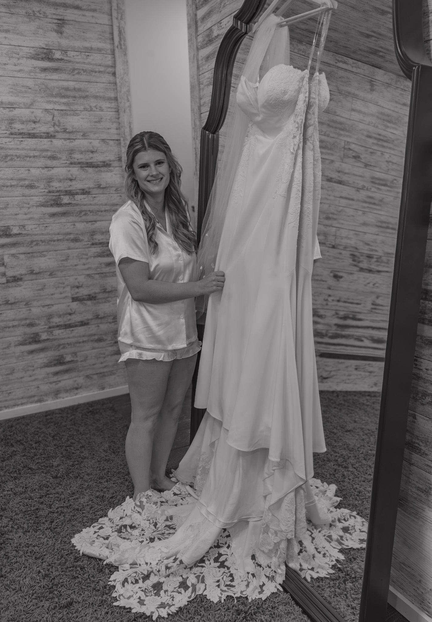 Black and white photo of a bride in pajamas smiling beside her elegant lace wedding gown hanging on display.