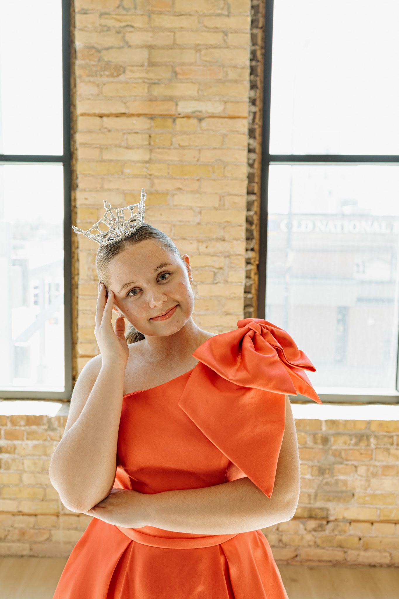 Young woman in orange gown and silver crown poses gracefully in a brick-walled studio.