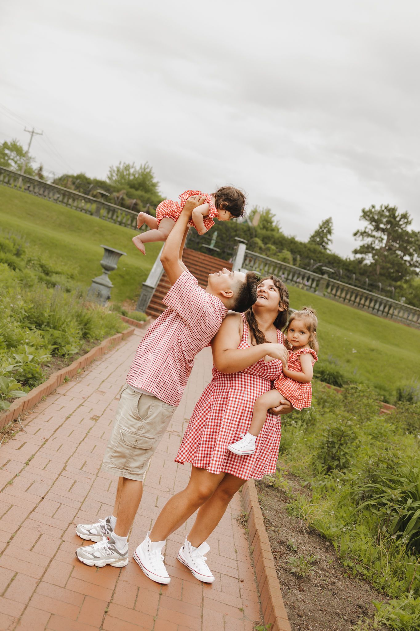 Joyful family in matching red outfits playing on a brick path, father lifting baby in the air.
