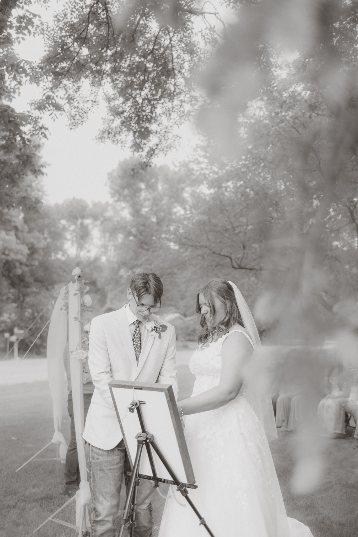Black and white photo of bride and groom emotional during outdoor wedding unity ceremony at easel.