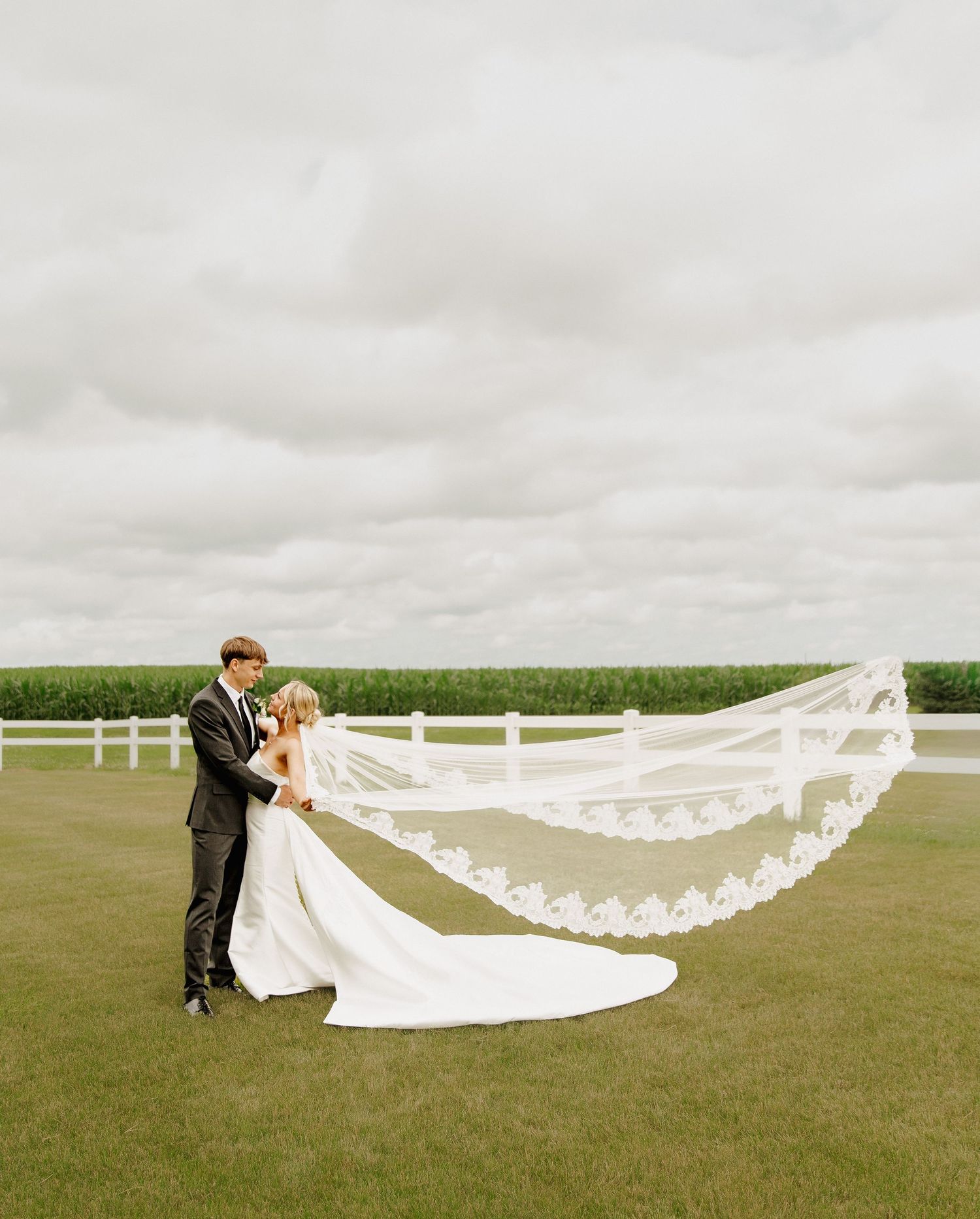 Bride and groom embrace on green lawn as cathedral lace veil flows dramatically in the wind at outdoor wedding.
