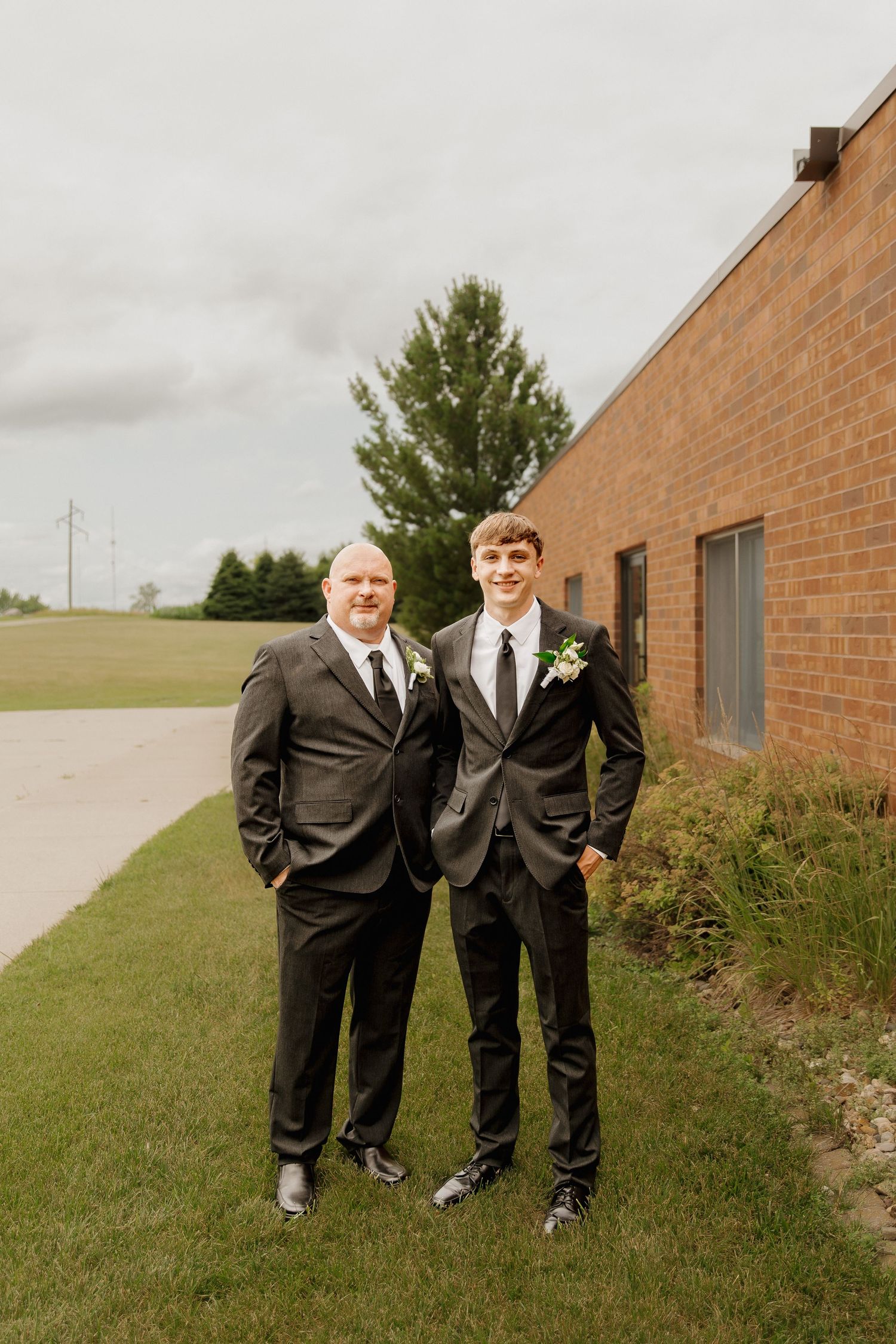 Two men in dark suits with boutonnieres posing outdoors beside brick building on wedding day.