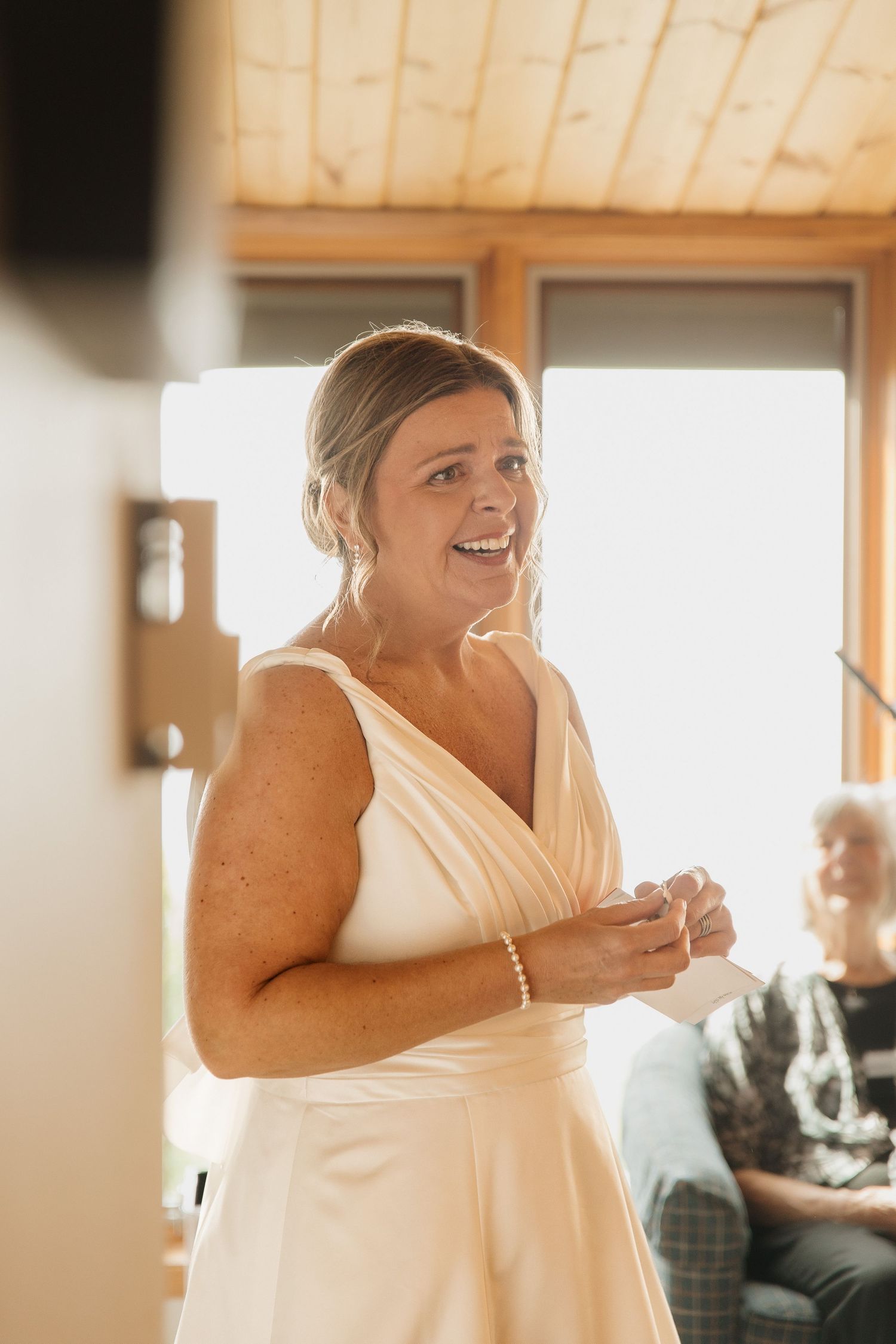 Smiling bride in elegant white V-neck wedding gown holding a note indoors on her wedding day.