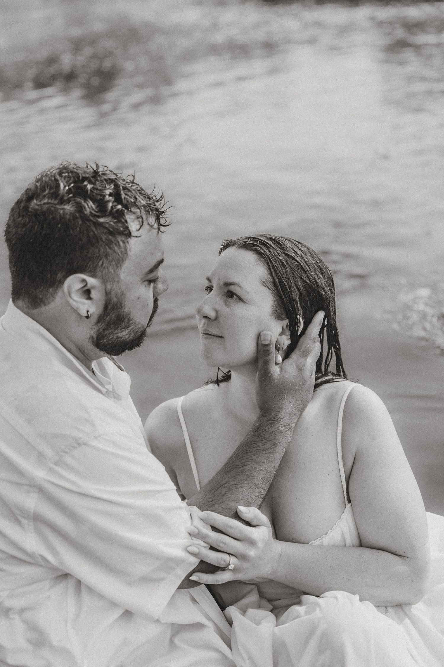 Black and white photo of a couple gazing at each other near water, man tenderly touching womans face.