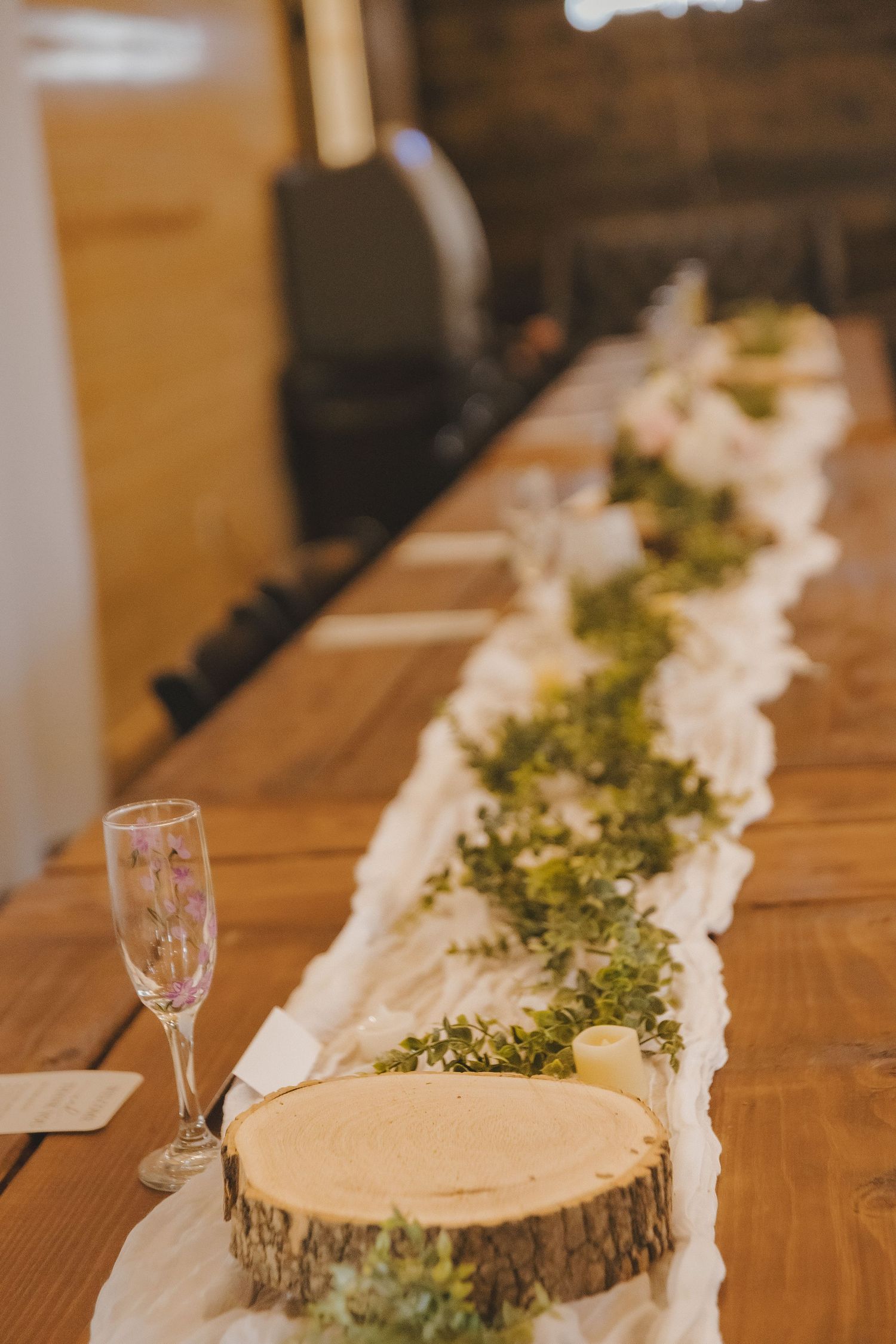 Rustic wedding head table with wood slice centerpieces, greenery garland, white runner, and champagne flute in a barn venue.