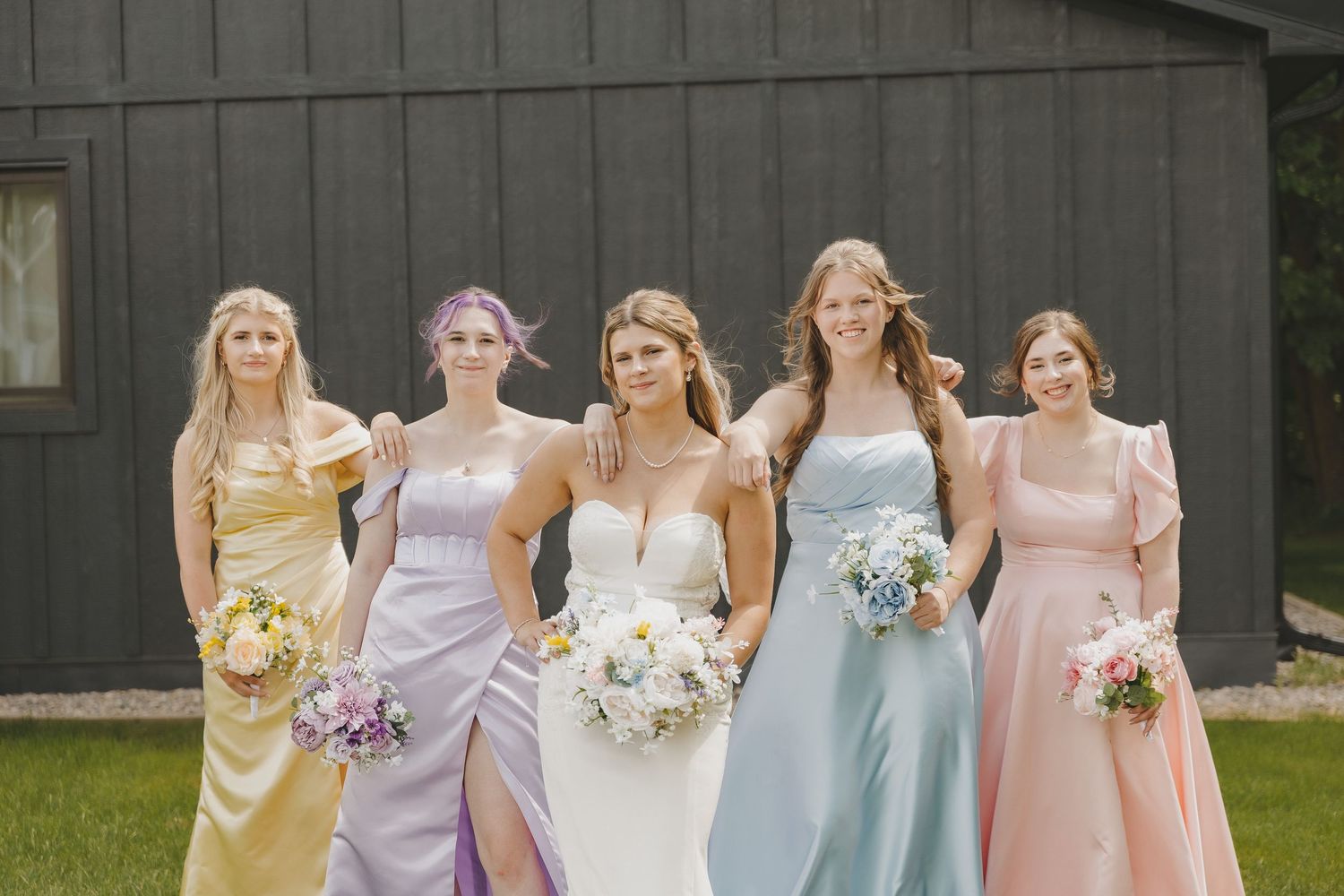 Bride in white gown poses with four bridesmaids in pastel mismatched dresses holding bouquets outside a dark barn.