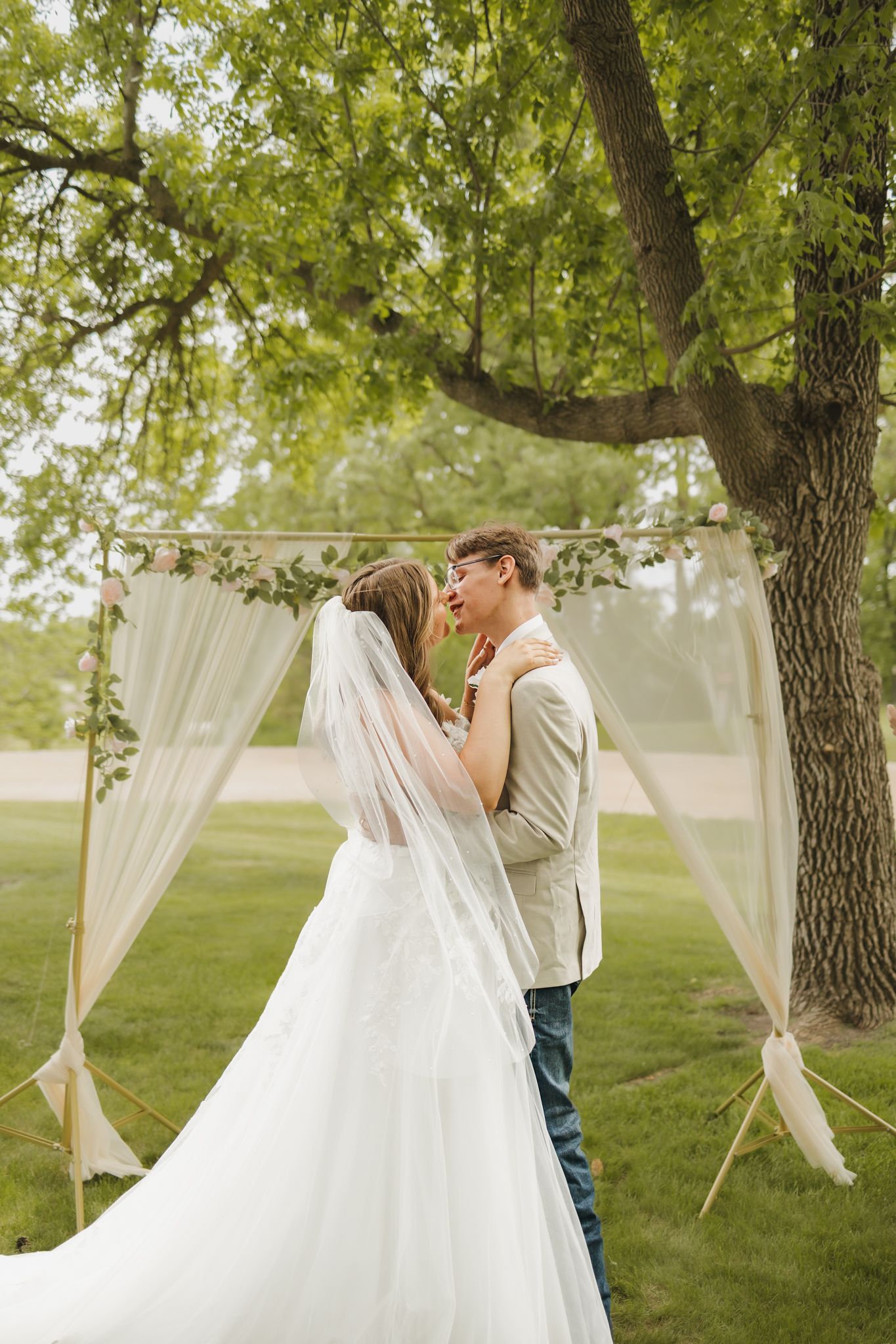 Bride and groom share first kiss under a draped outdoor wedding arch surrounded by lush green trees.
