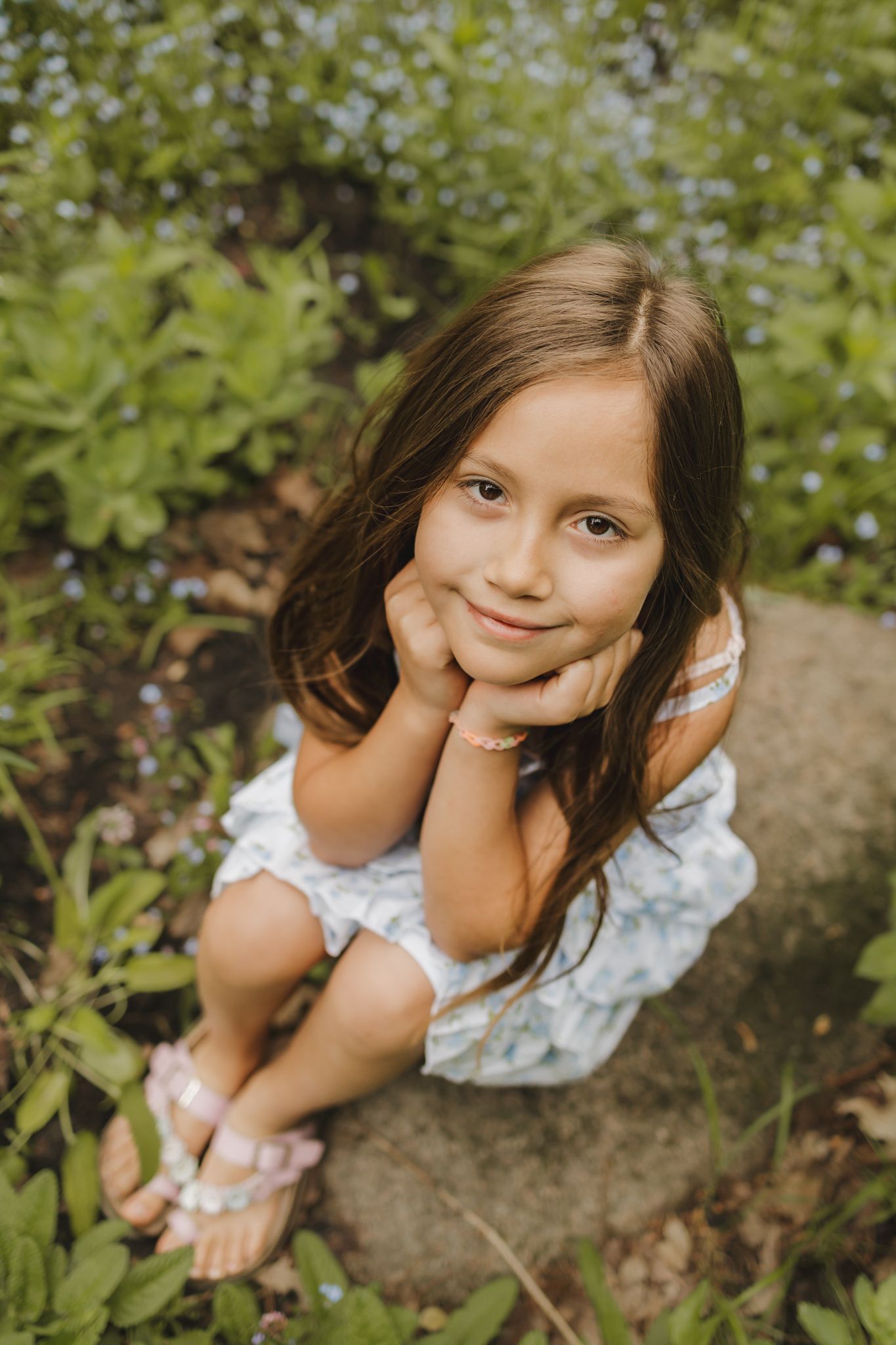 Adorable young girl with long brown hair sitting on a rock among garden flowers, smiling sweetly.