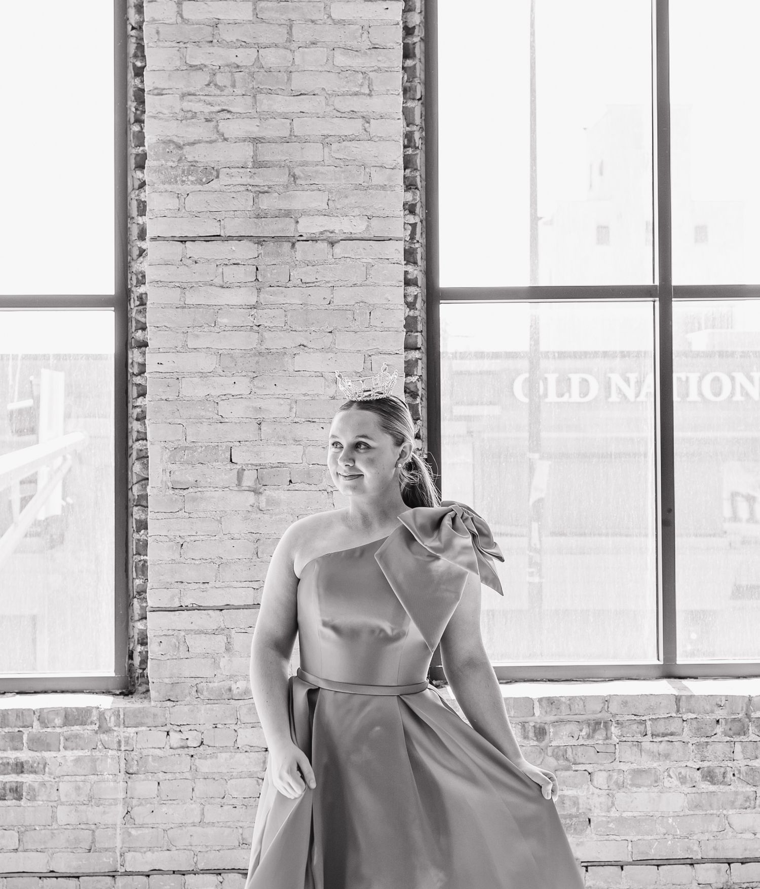 Woman in elegant one-shoulder gown poses gracefully in black and white photo near large industrial windows.