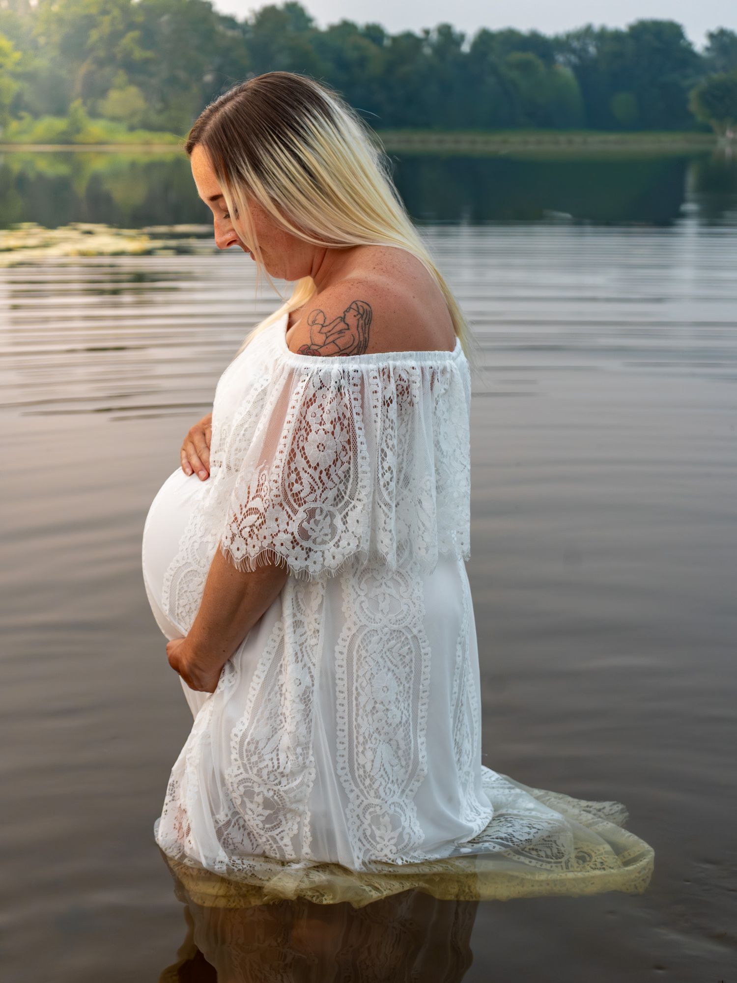 Artistic maternity portraits taken in a white lace dress at the water's edge.