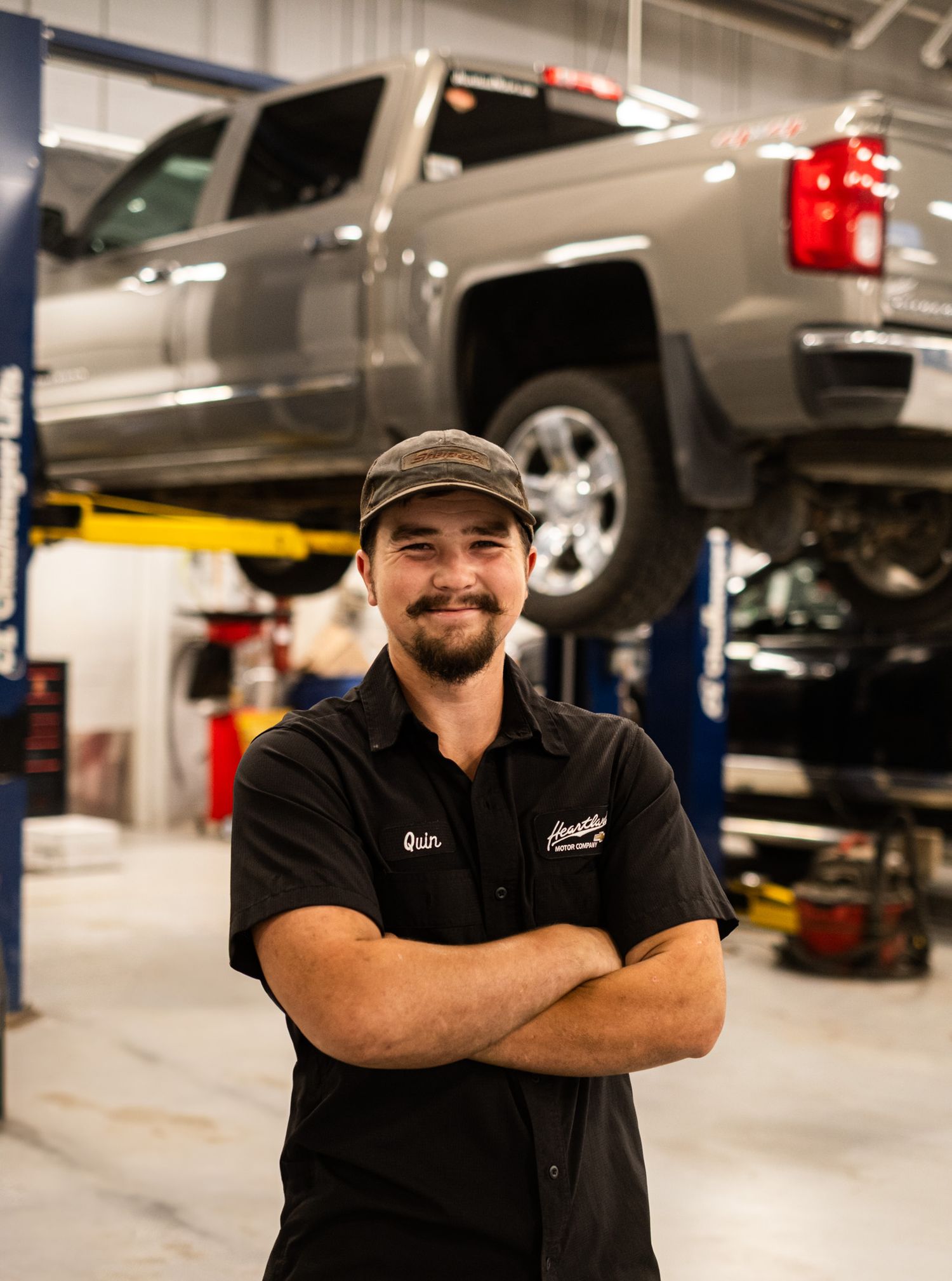 Auto technician in black uniform standing with crossed arms in front of elevated pickup truck.