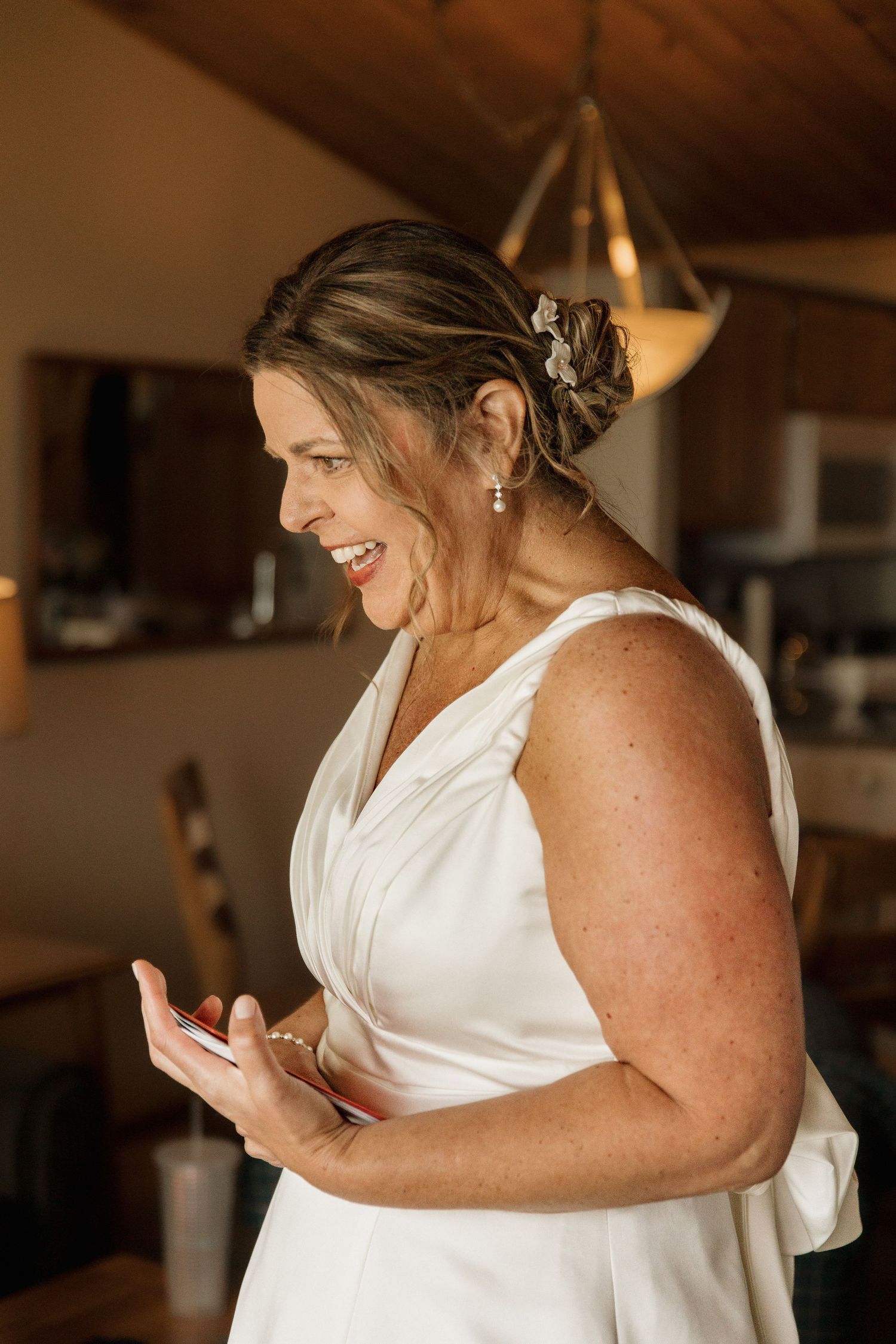 Smiling bride in elegant white sleeveless wedding gown reading a note indoors with warm golden lighting.