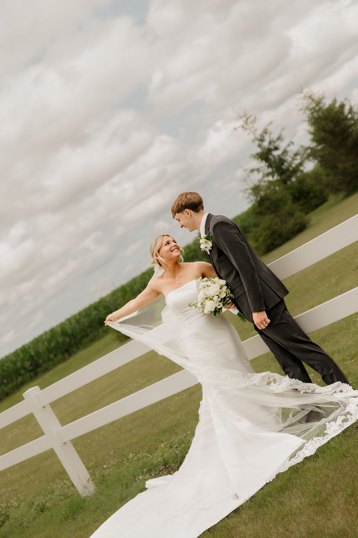 Bride and groom share a romantic moment by a white fence on a green farm landscape on their wedding day.