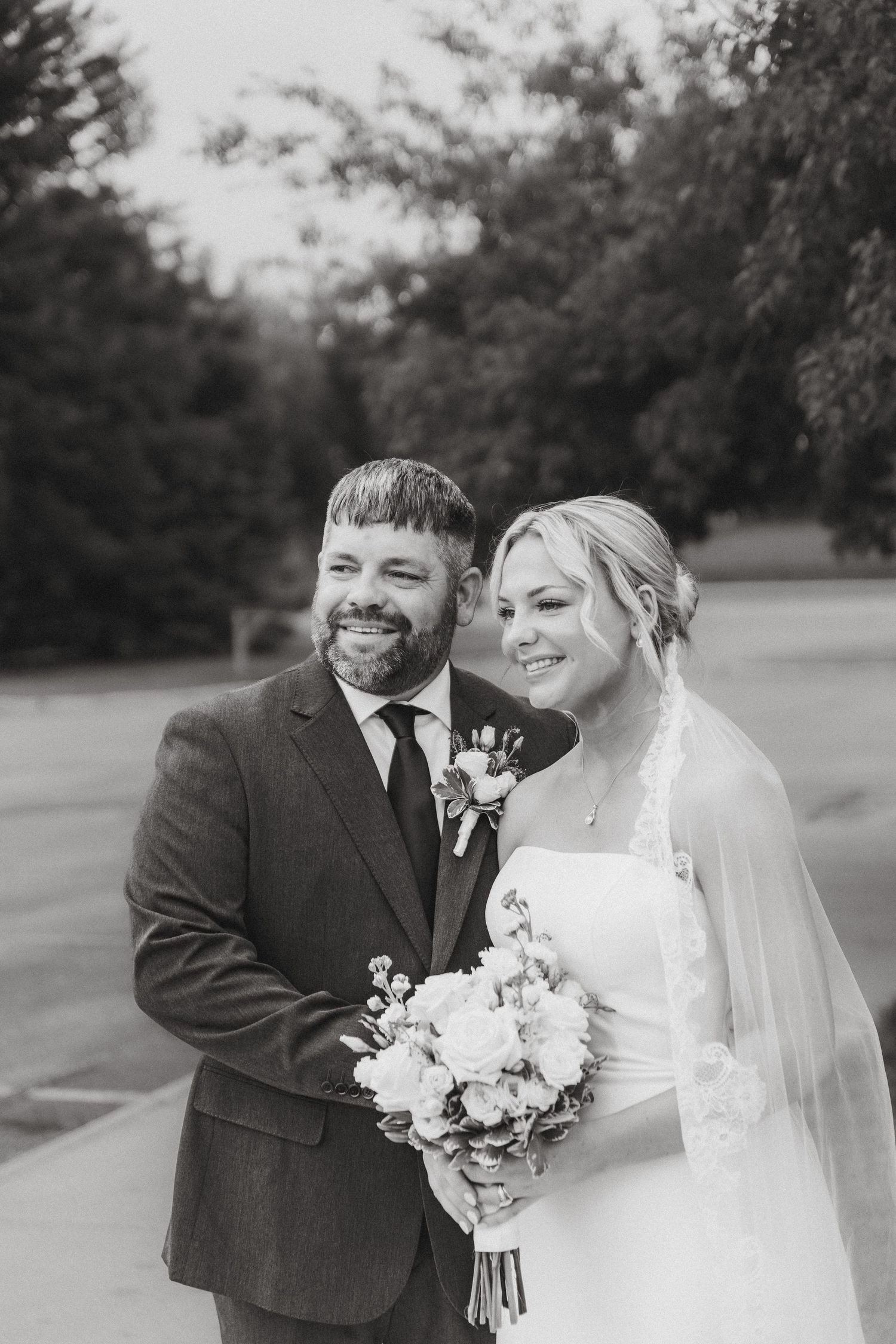 Black and white portrait of smiling bride and groom holding bouquet outdoors on wedding day.