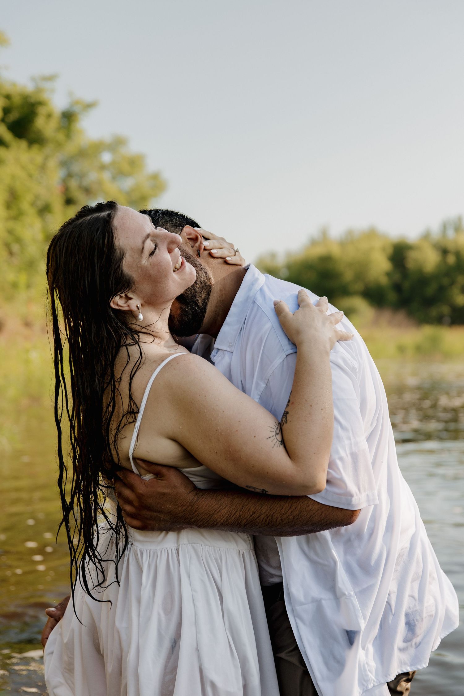 A couple in white clothing embraces romantically near a river, the woman with wet hair tilting her head back joyfully.