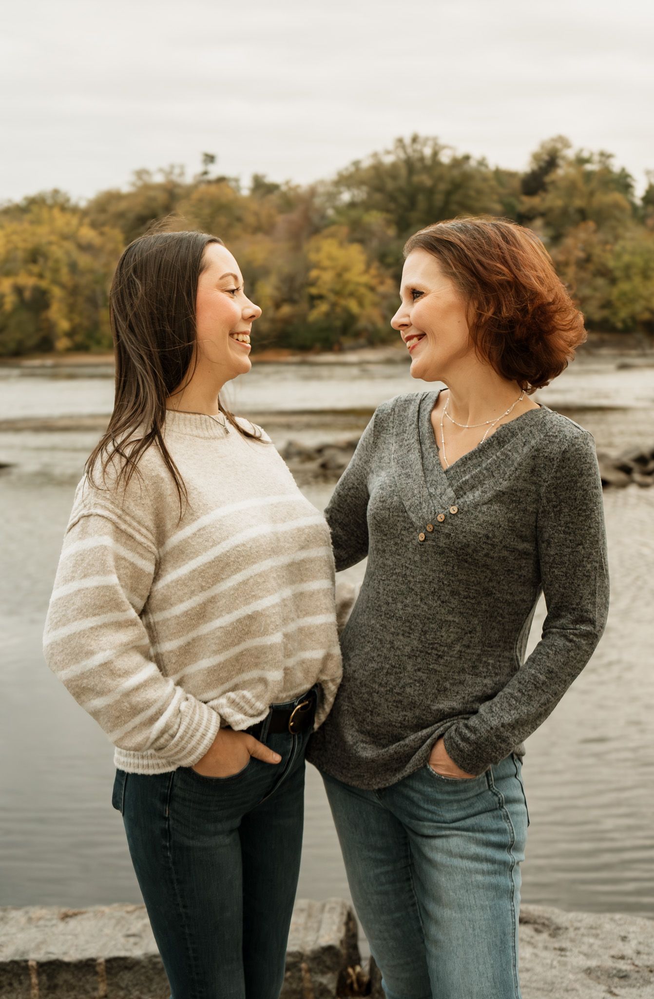 Two people in sweaters laugh together during an outdoor photoshoot by the water.