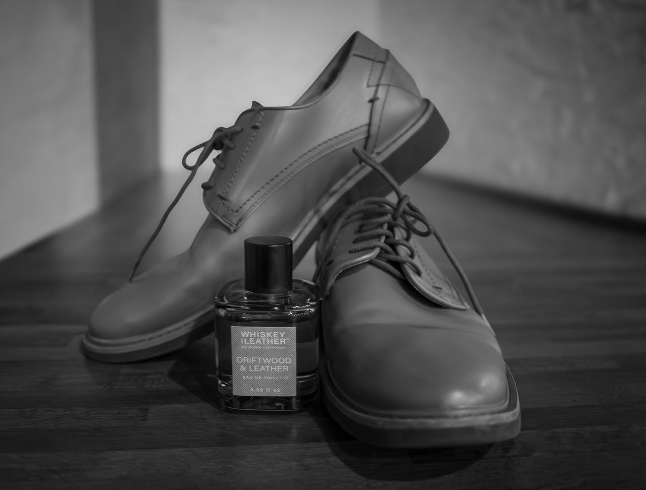 A pair of formal black leather dress shoes with a small bottle of cologne on a wooden floor.