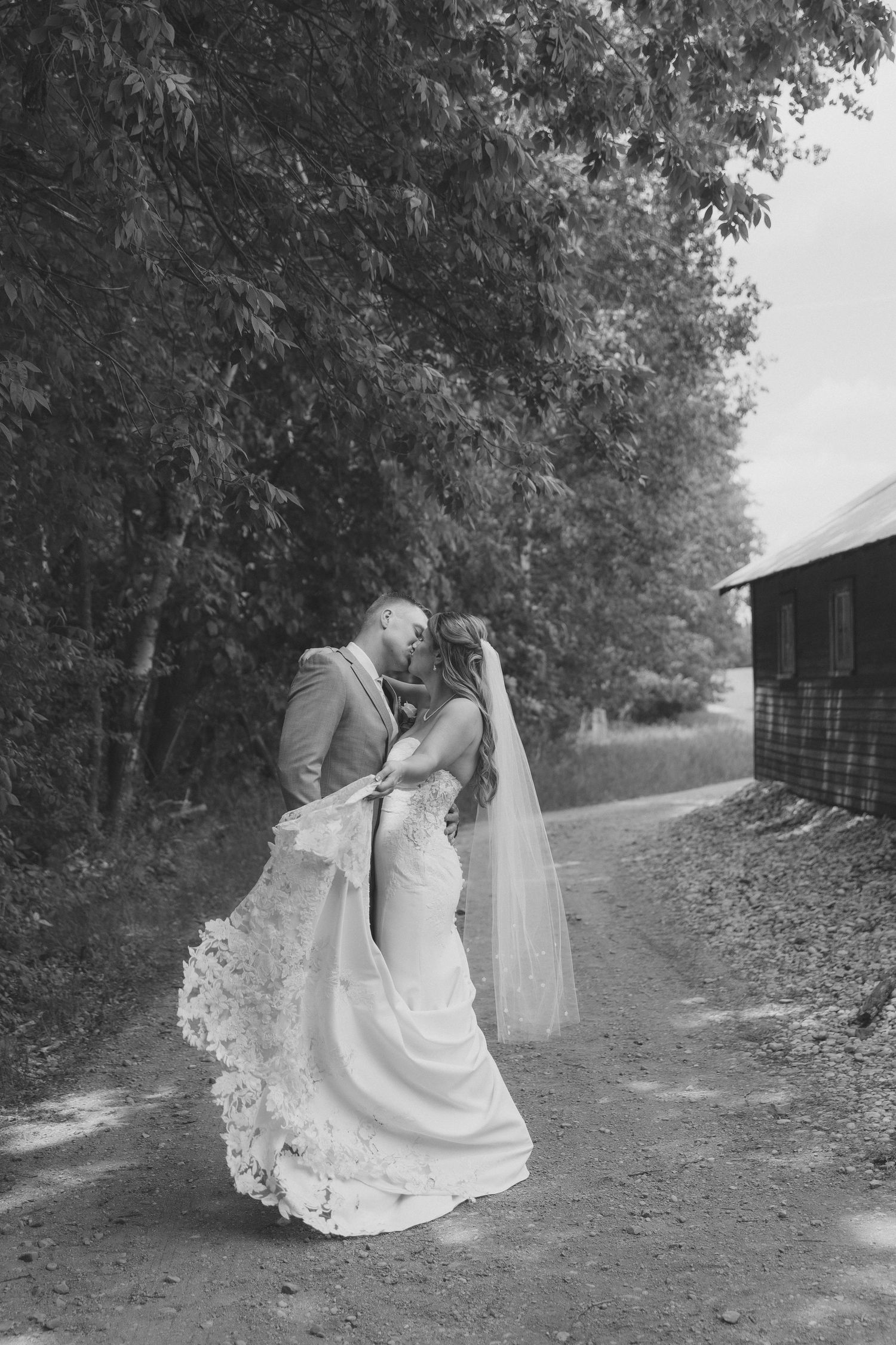 Black and white photo of a bride and groom sharing a romantic kiss on a rustic path surrounded by trees.