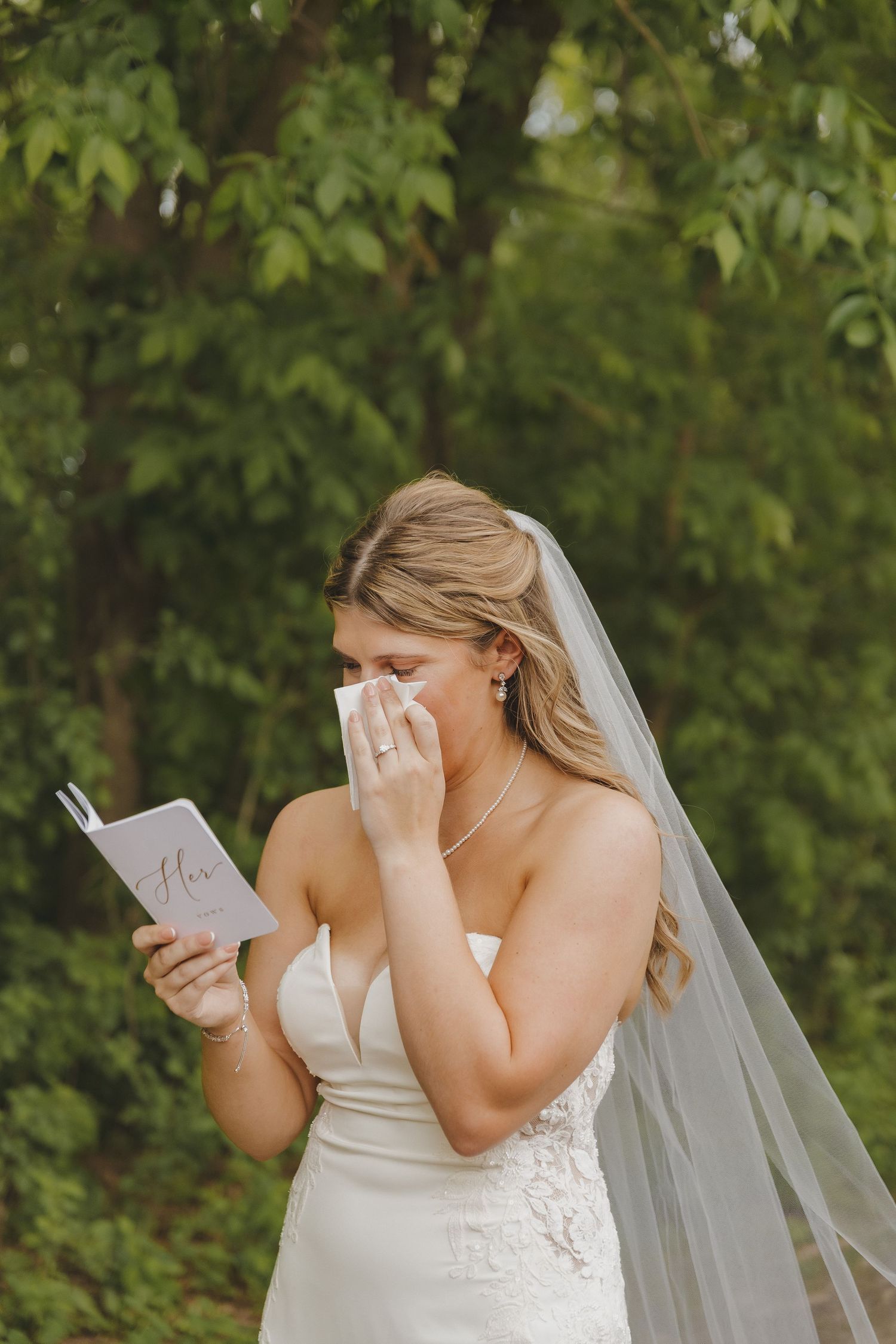 Emotional bride in white gown and veil crying while reading wedding vows card outdoors.