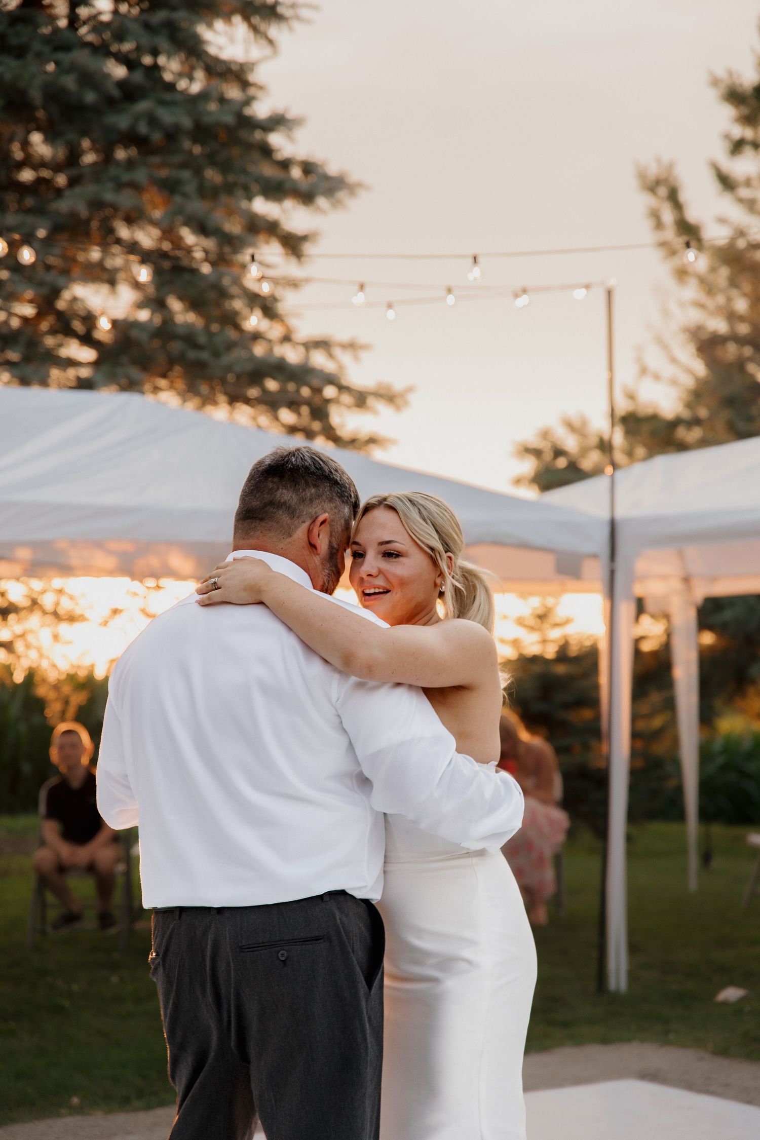 Bride in white dress shares joyful first dance outdoors at sunset under string lights at reception.