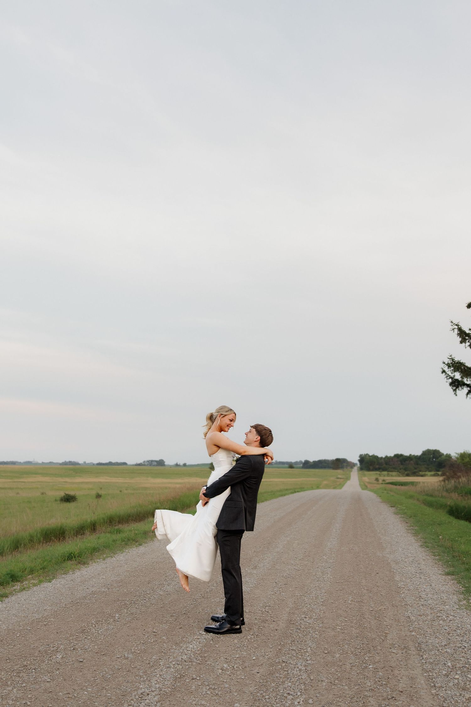Groom lifting bride in white dress on rural gravel road surrounded by open green fields.