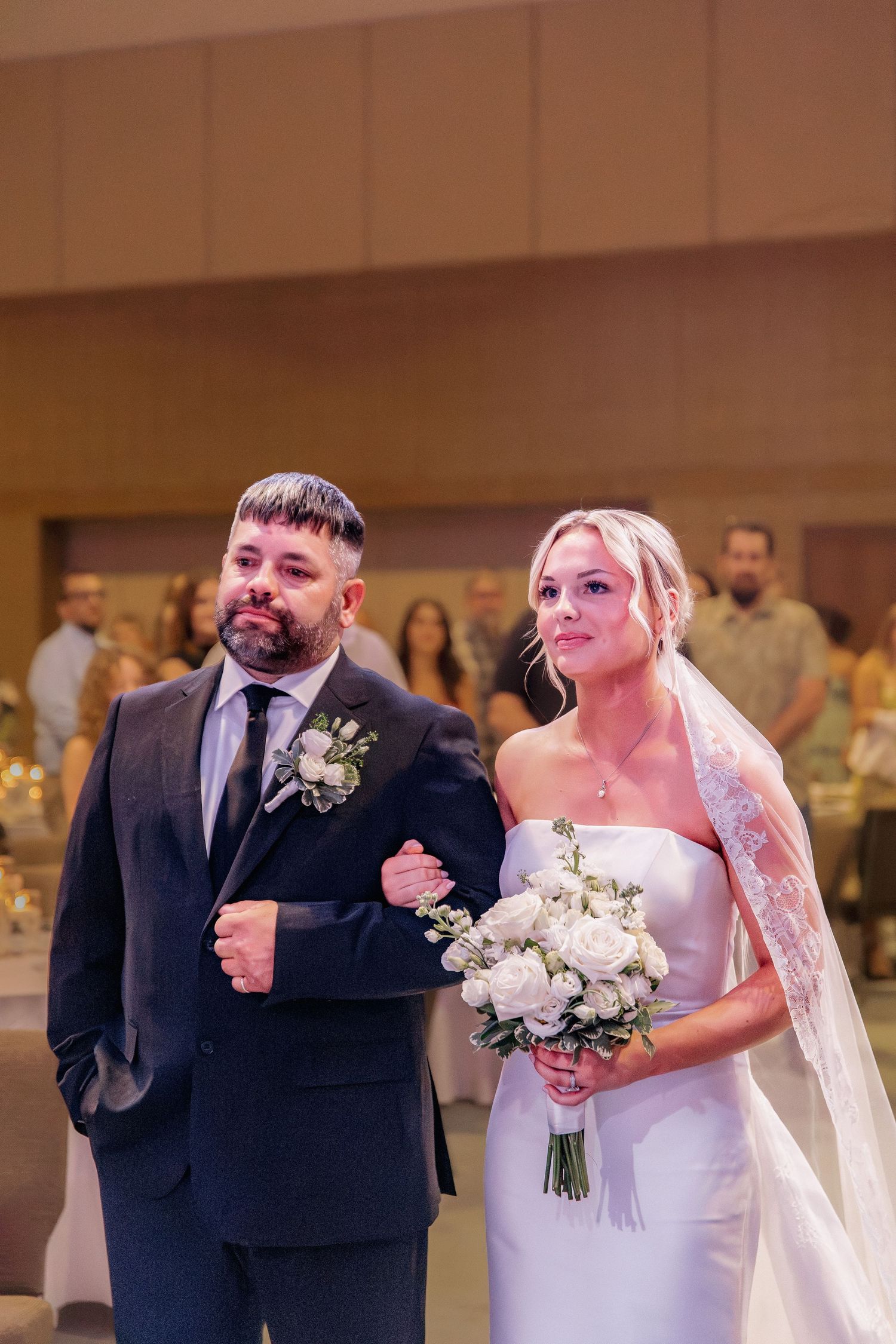 Bride in white gown with veil and groom in dark suit walking down aisle at indoor wedding ceremony.