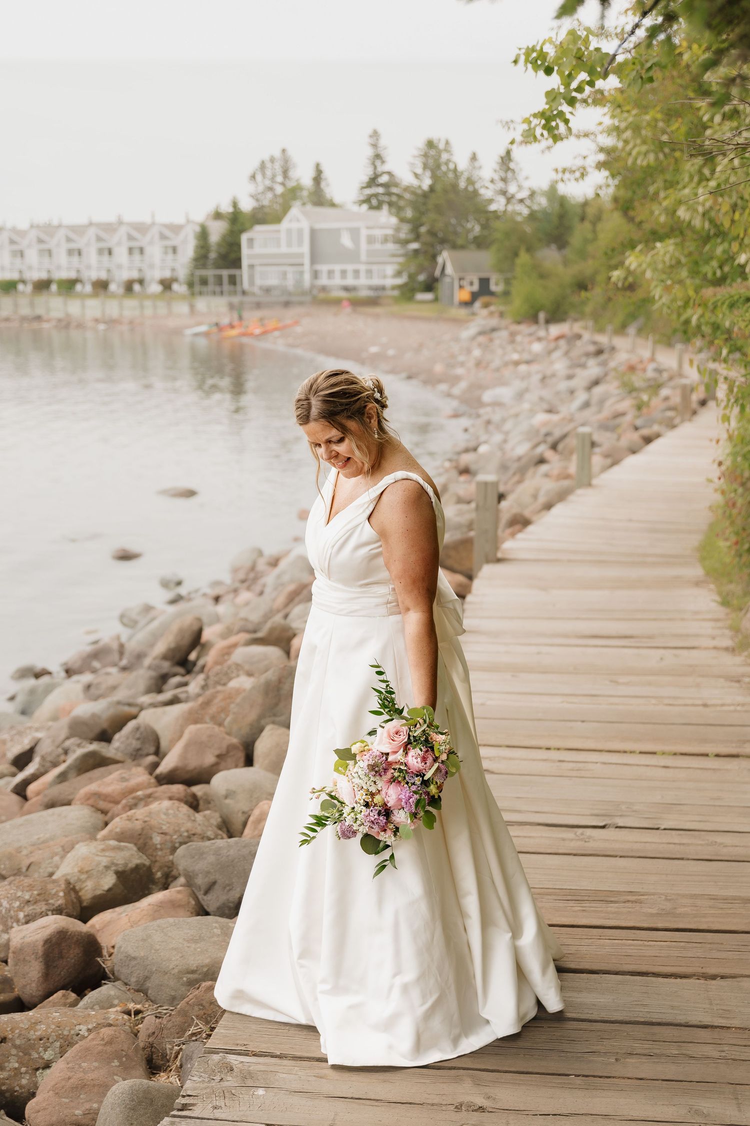 Bride in a white gown holding a bouquet walks on a wooden dock beside a rocky lakeshore.