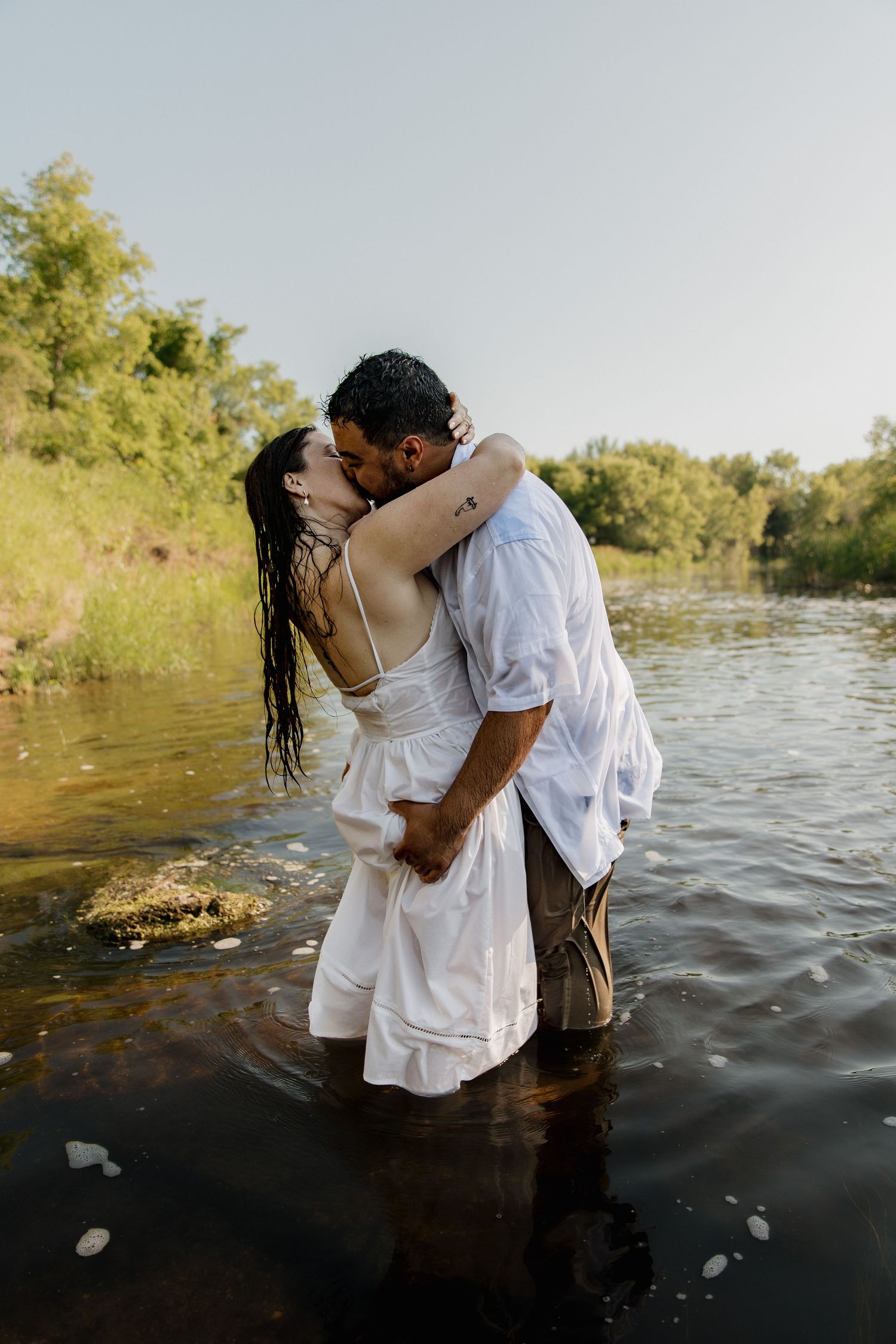 Couple sharing a romantic kiss while standing in a shallow river, woman in white dress, lush green trees in background.