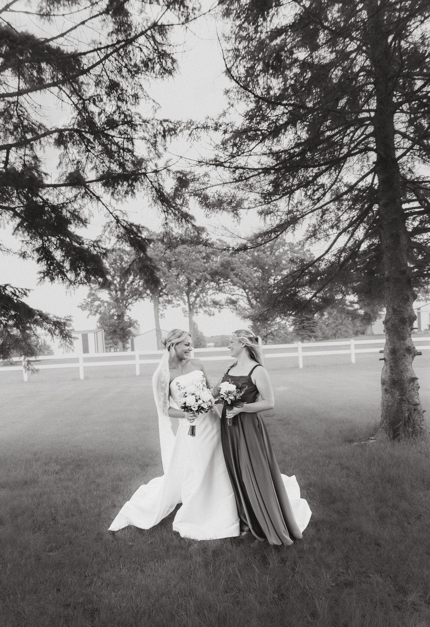 Black and white photo of bride and bridesmaid sharing a moment under pine trees by white fence.
