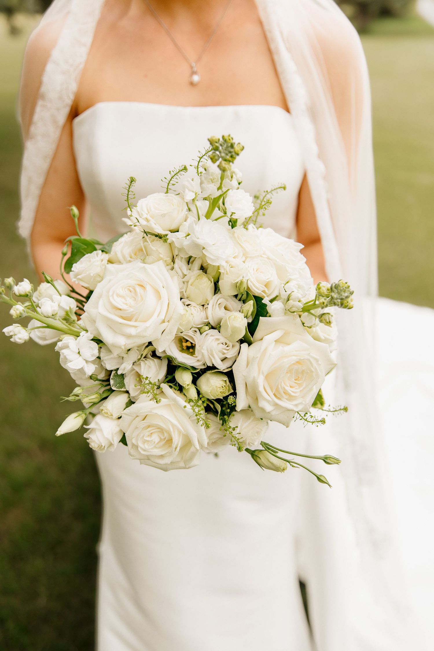 Bride holding a lush white rose wedding bouquet with greenery accents in a strapless gown.