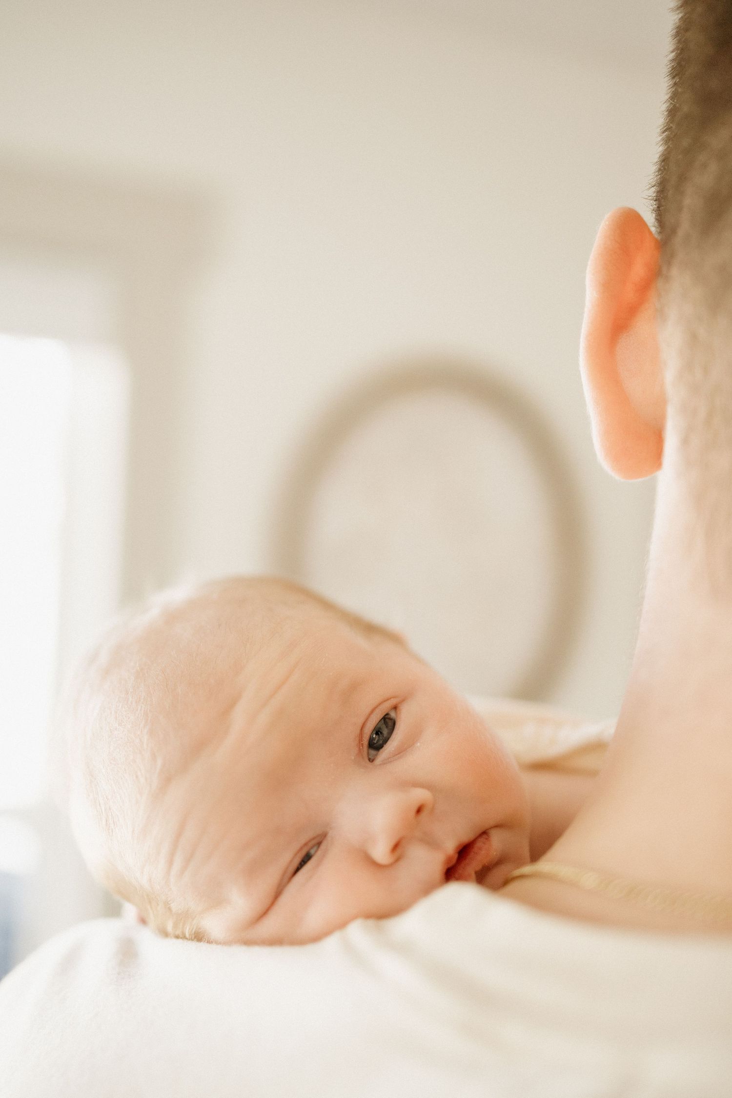 Newborn baby resting peacefully on father's shoulder, looking at camera in bright white nursery.